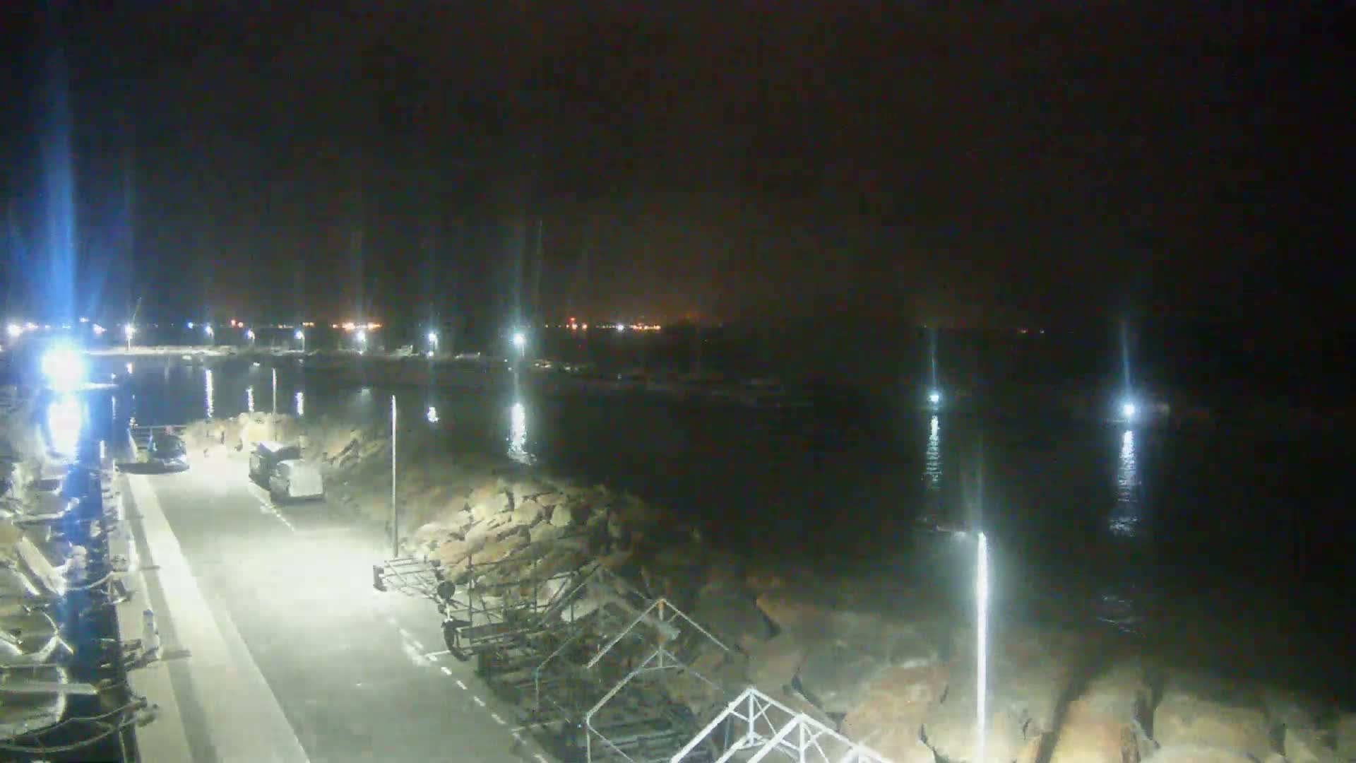 A nighttime view of a harbor, with boats moored and lights illuminating the calm water and a paved walkway.