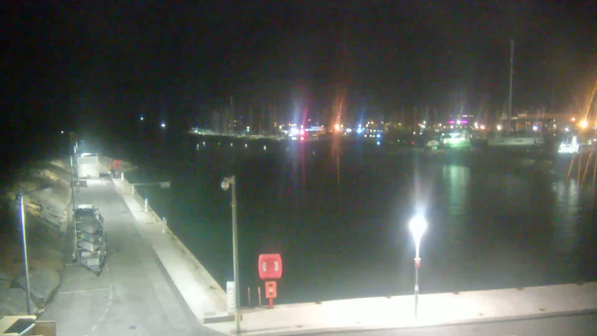 A nighttime view of a harbor with docked boats, lit by streetlights, and a paved walkway extending from the foreground.