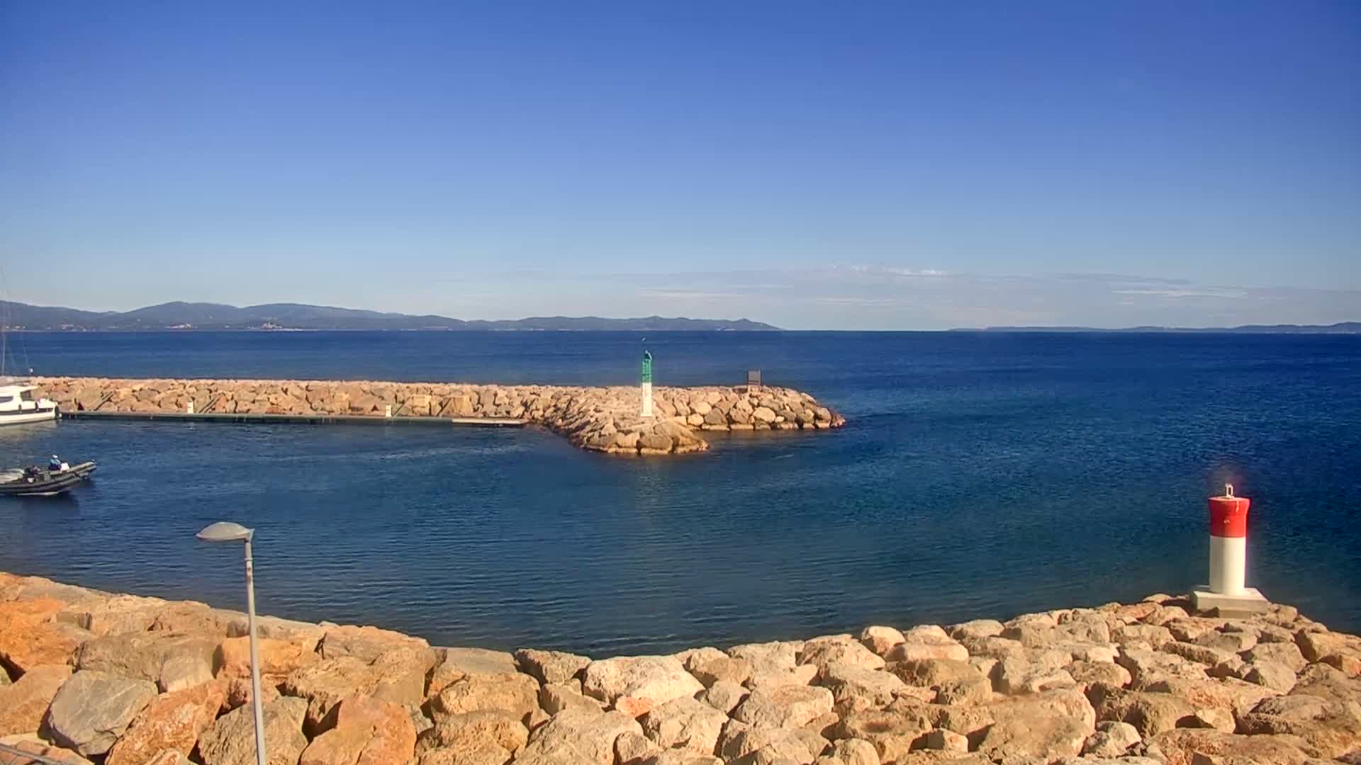 A calm, blue sea stretches to a distant mountain range under a clear blue sky, with a rocky breakwater featuring a small lighthouse and a few moored boats.