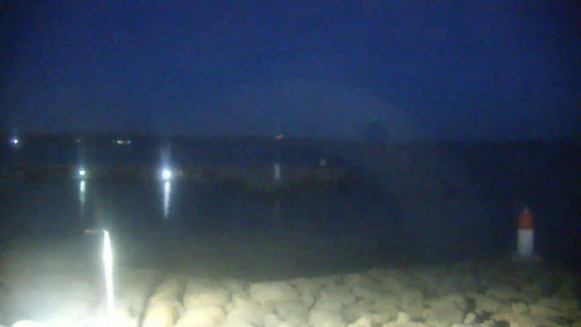 A dark, nighttime scene shows a body of water with a rocky shoreline, a small pier, and several lights reflecting on the water.
