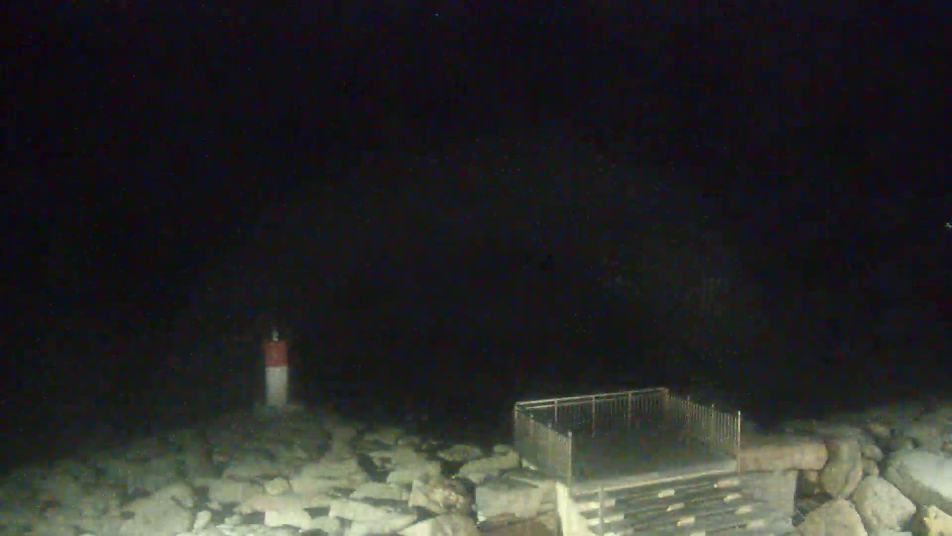 A nighttime, low-light image shows a rocky shoreline with a small fenced-in structure and a navigational marker.