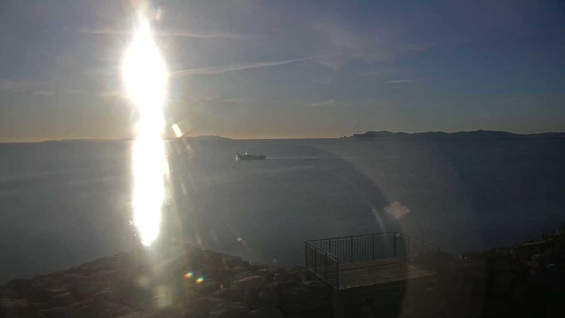 A small boat is visible on a calm body of water under a bright, sunny sky, viewed from a rocky outcrop with a small railing and platform.