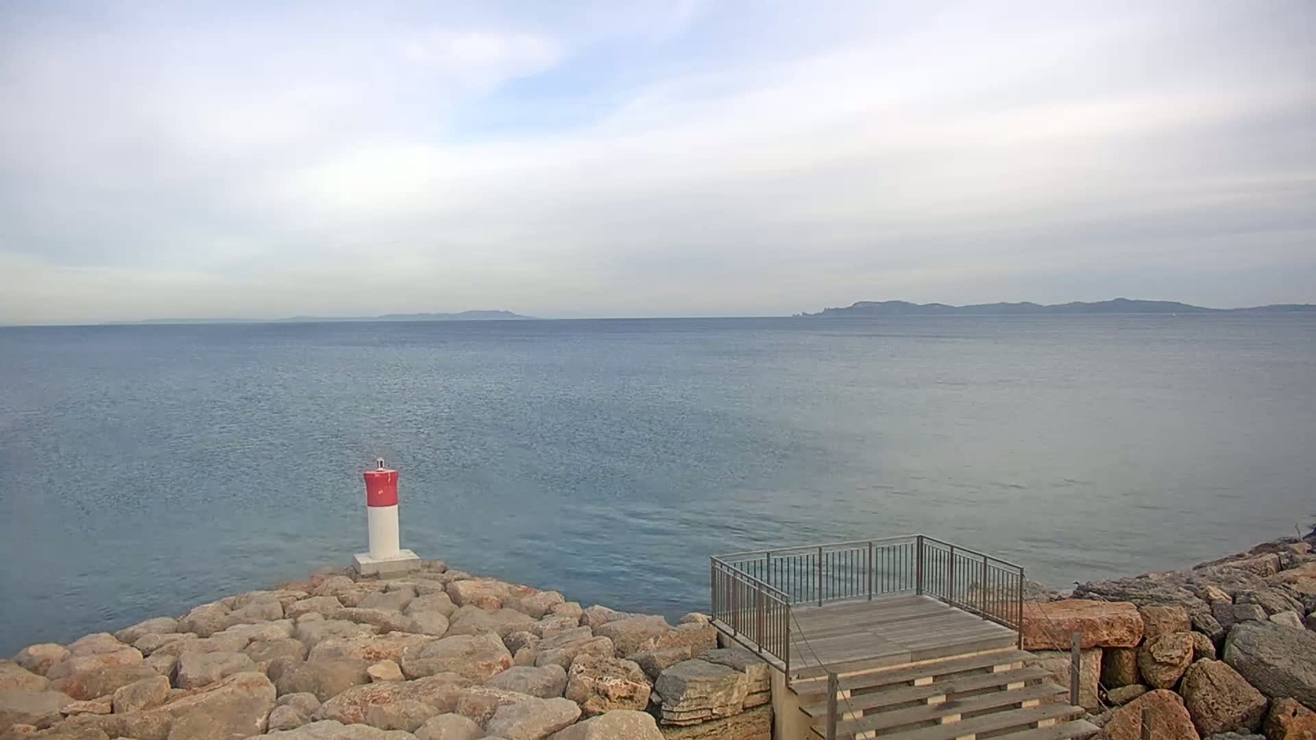 A rocky shoreline with a small lighthouse and a wooden platform with stairs overlooks a calm sea under a cloudy sky, with distant landmasses visible on the horizon.