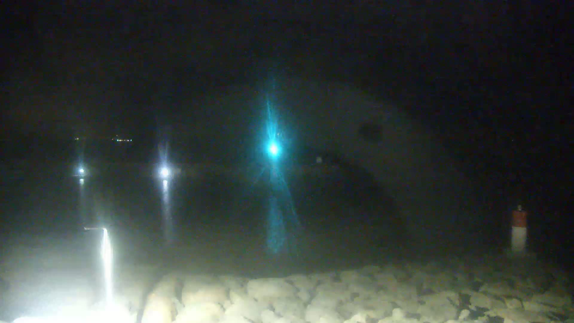 A nighttime scene shows a rocky shoreline with a body of water in the background, illuminated by several lights including a bright, teal light in the center.