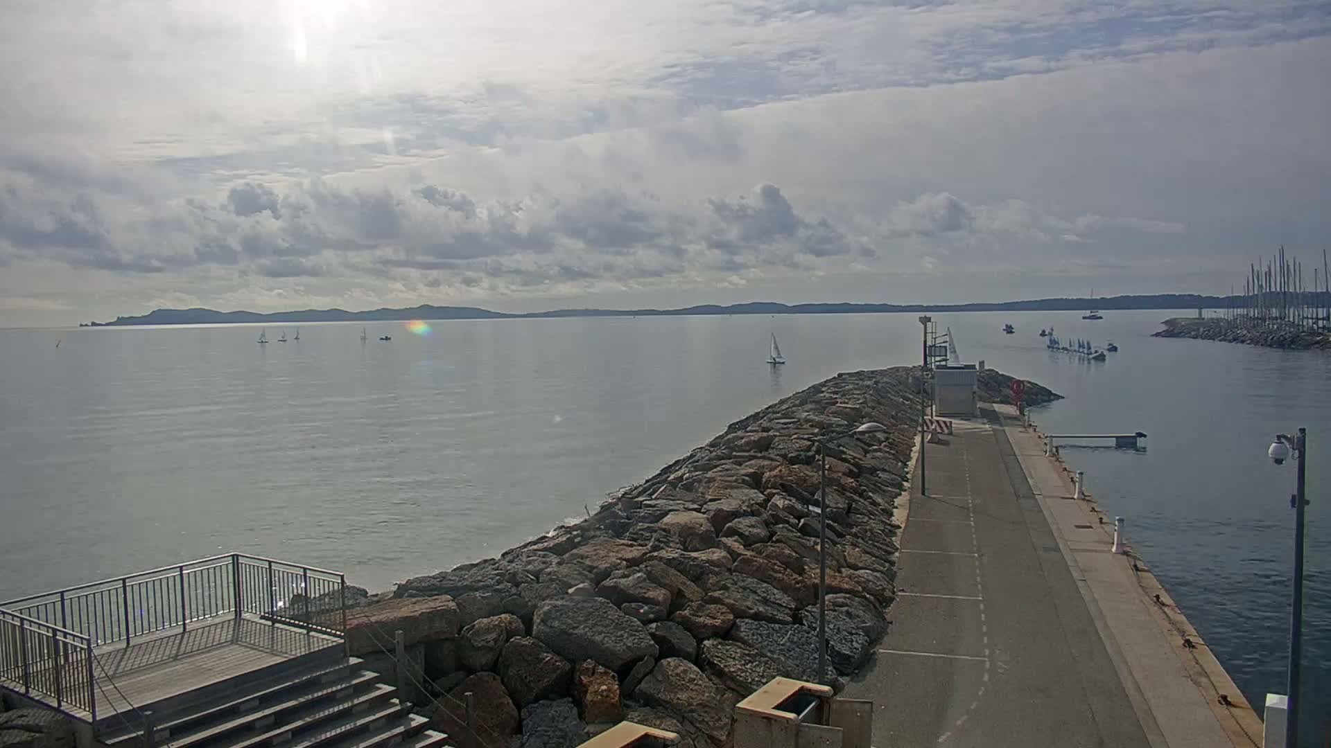 A rocky jetty extends into a calm sea under a partly cloudy sky, with several sailboats visible in the distance.