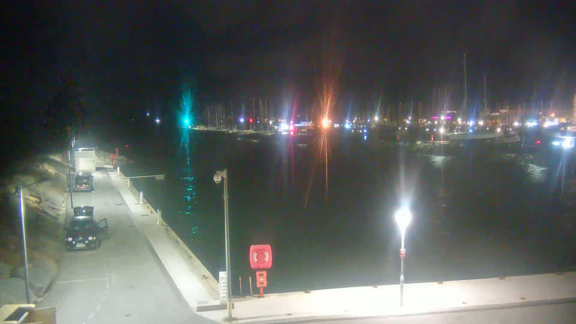 A nighttime view of a harbor filled with boats, a calm body of water, and a roadway with two parked cars.