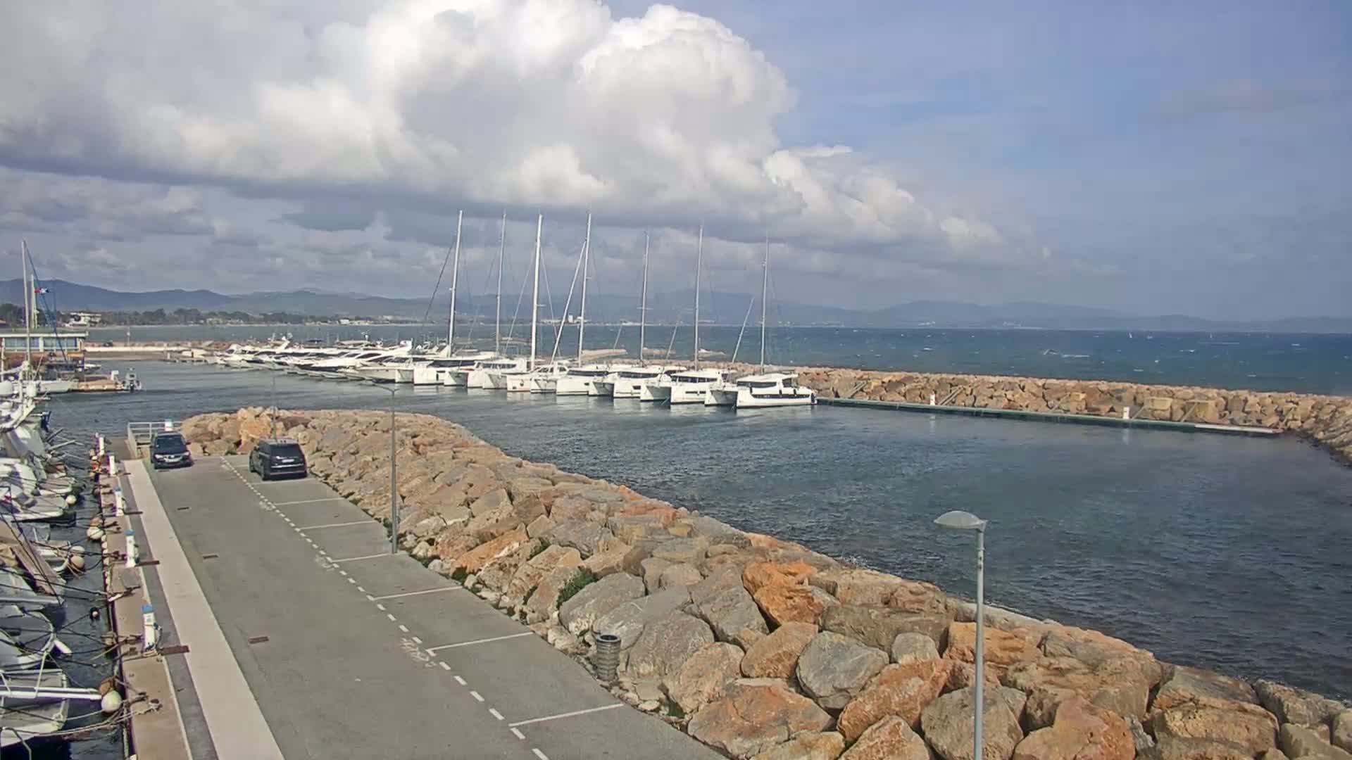 A marina filled with sailboats and catamarans is protected by a rock wall, overlooking a partly cloudy sky and a choppy sea.