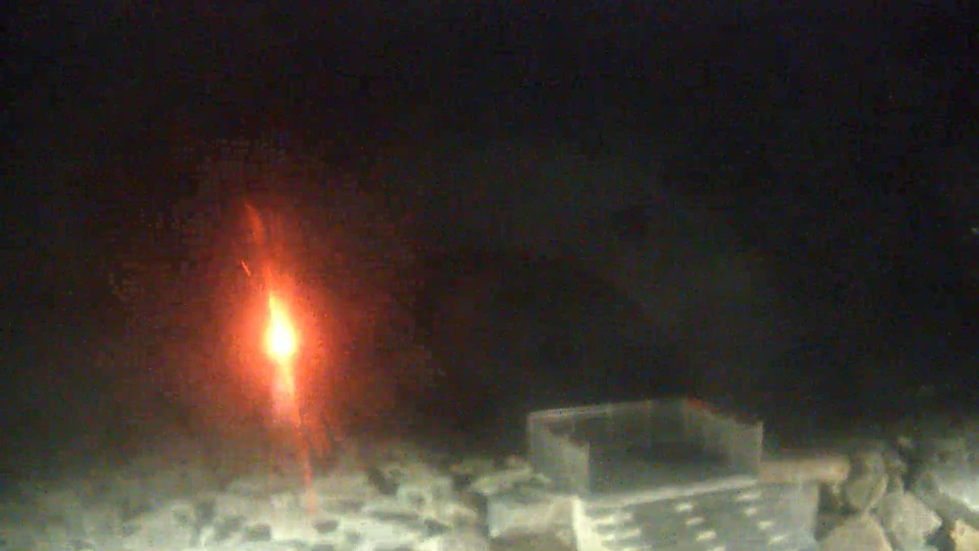 A tall, slender flame burns brightly amidst a pile of dark gray rocks and a small, rectangular metal container in a dark, enclosed space.