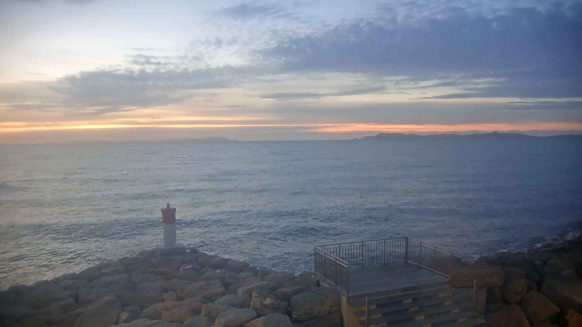 A rocky shoreline with a small lighthouse and viewing platform overlooks a calm sea under a partly cloudy sunset sky.