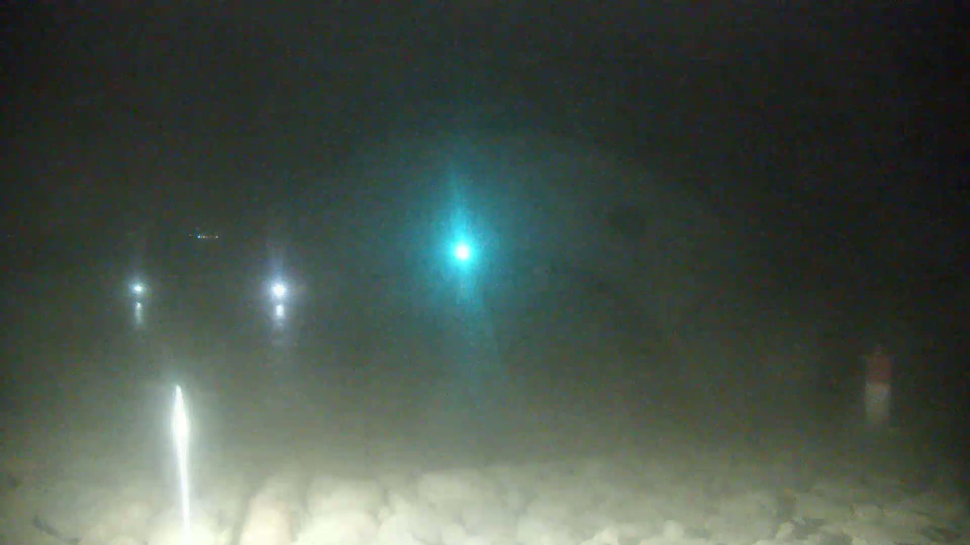 At night, in hazy conditions, several lights are visible in the distance along a rocky, possibly beach-like area.