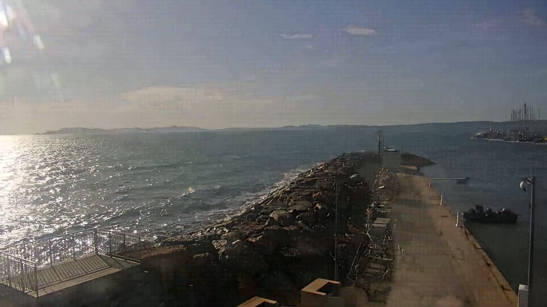 A rocky breakwater extends into a moderately wavy sea under a mostly sunny sky, with distant land visible on the horizon.