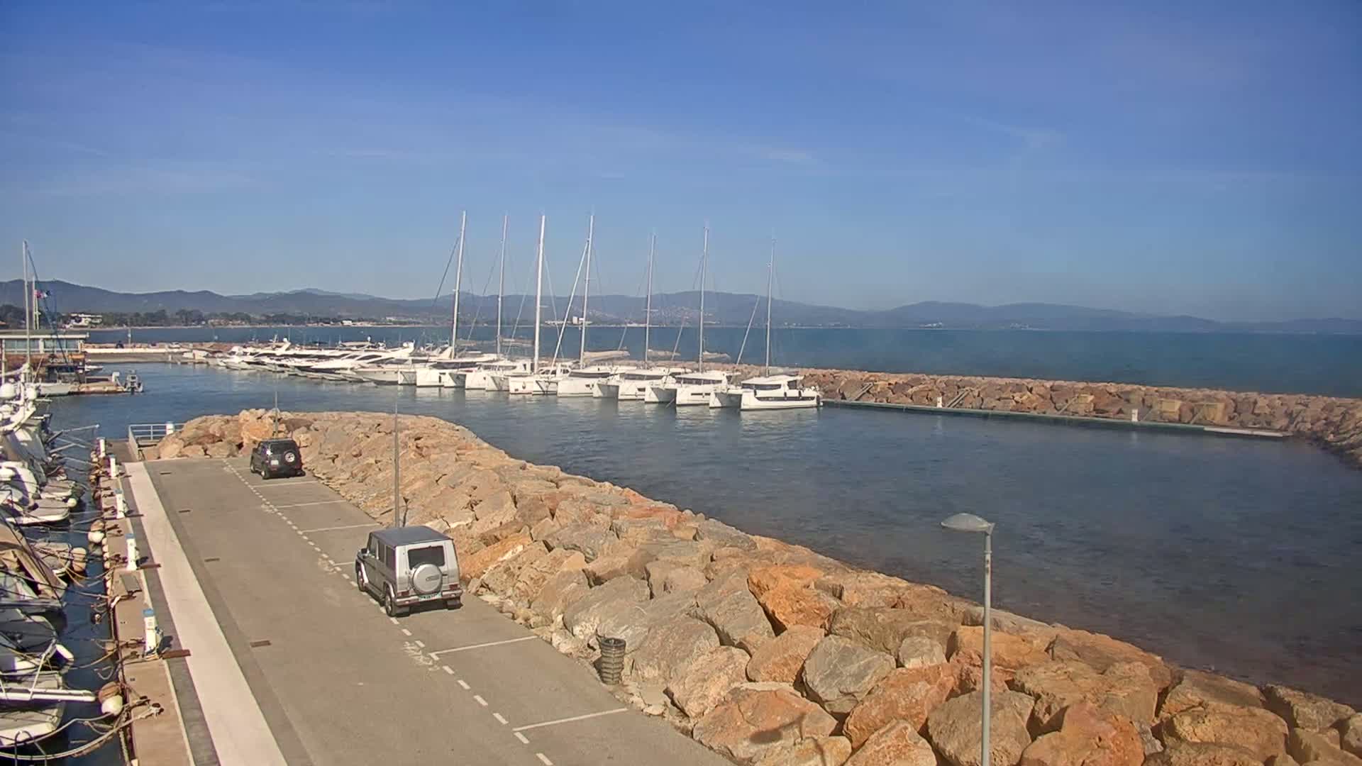 A marina with numerous sailboats and two cars parked on a paved area next to a rocky seawall, under a clear blue sky.