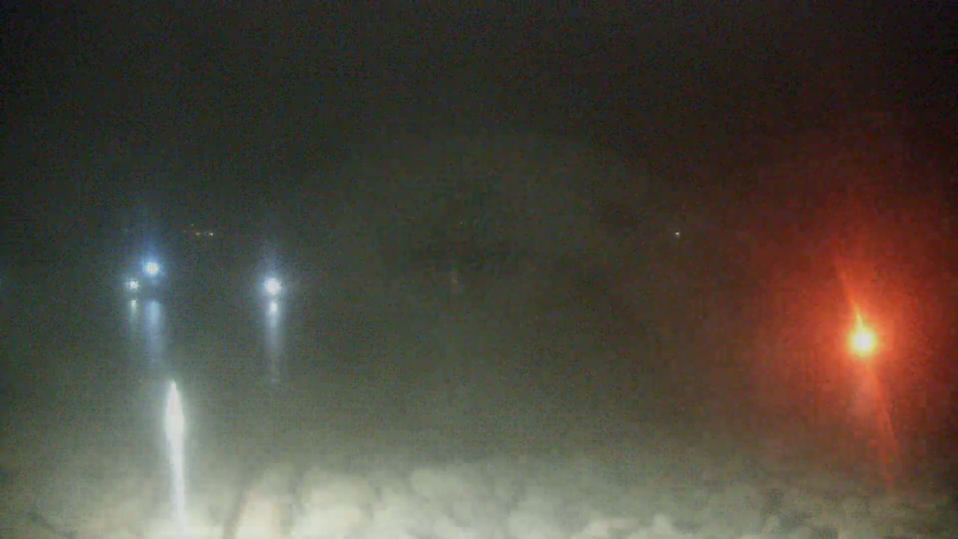 Three sets of vehicle headlights are visible through a foggy, dark nighttime scene, with a bright orange light appearing in the distance to the right.