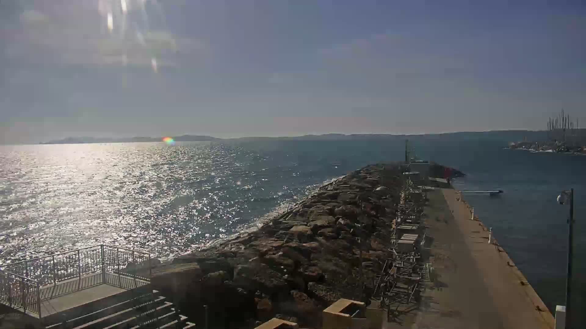 A rocky breakwater extends into a sunlit, slightly choppy sea, with distant landmasses visible on a clear day.