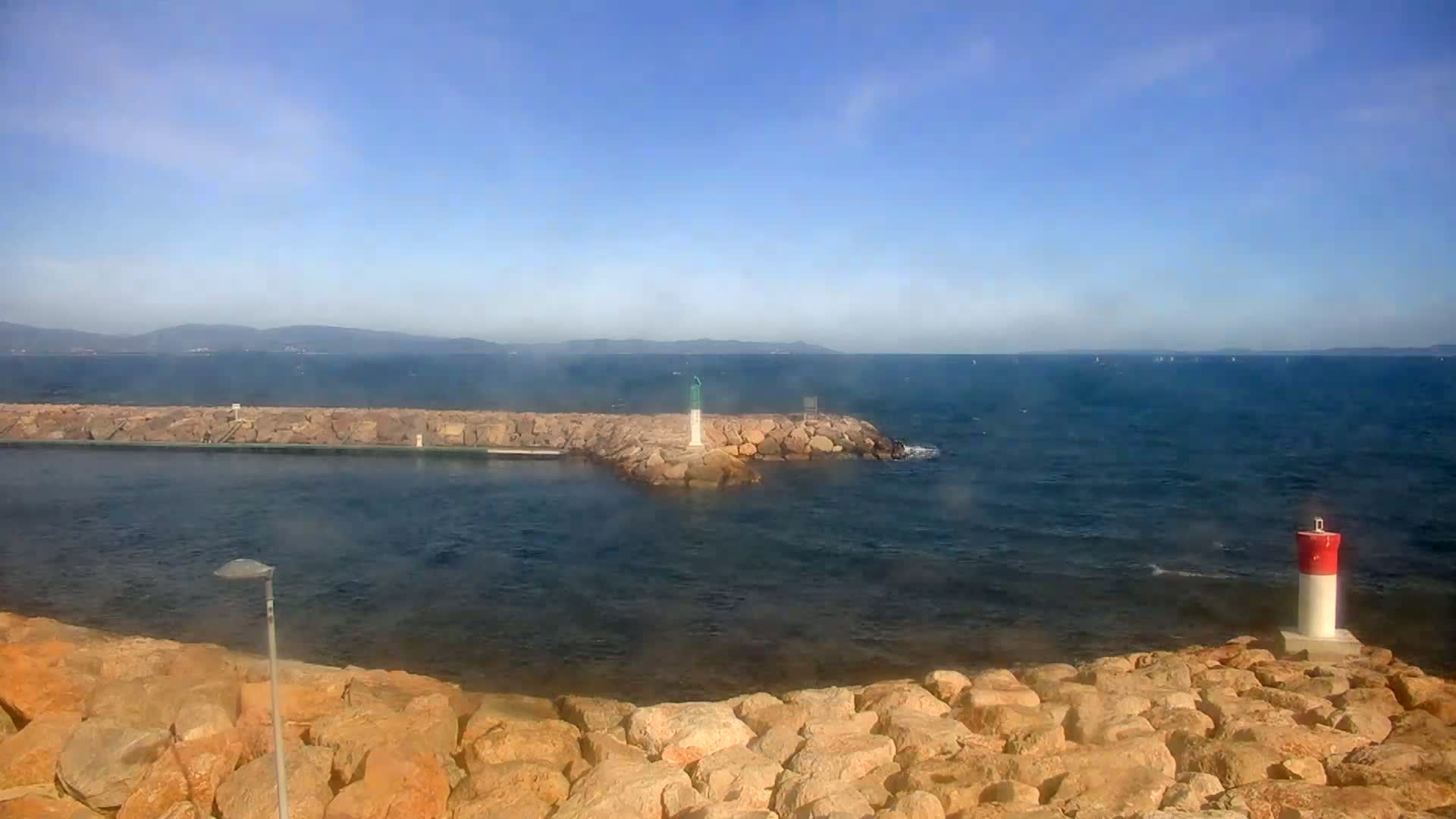 A rocky coastline with two small lighthouses, under a mostly clear blue sky, overlooks a calm sea with distant mountains visible on the horizon.
