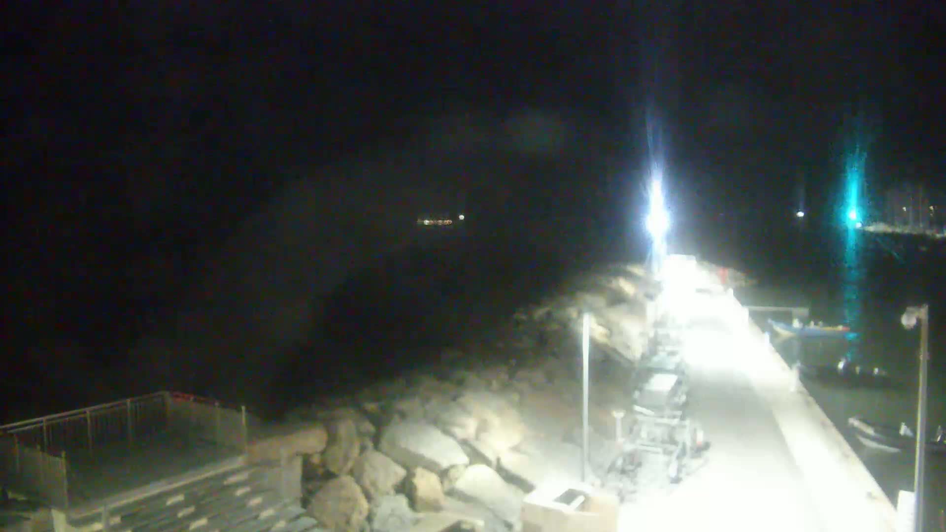 A nighttime view of a lit concrete pier extending into dark water, bordered by a rocky shoreline.