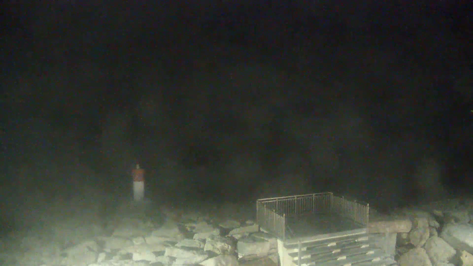 A dark, foggy night scene shows a metal staircase leading to a platform next to a rocky shoreline with a small marker in the distance.