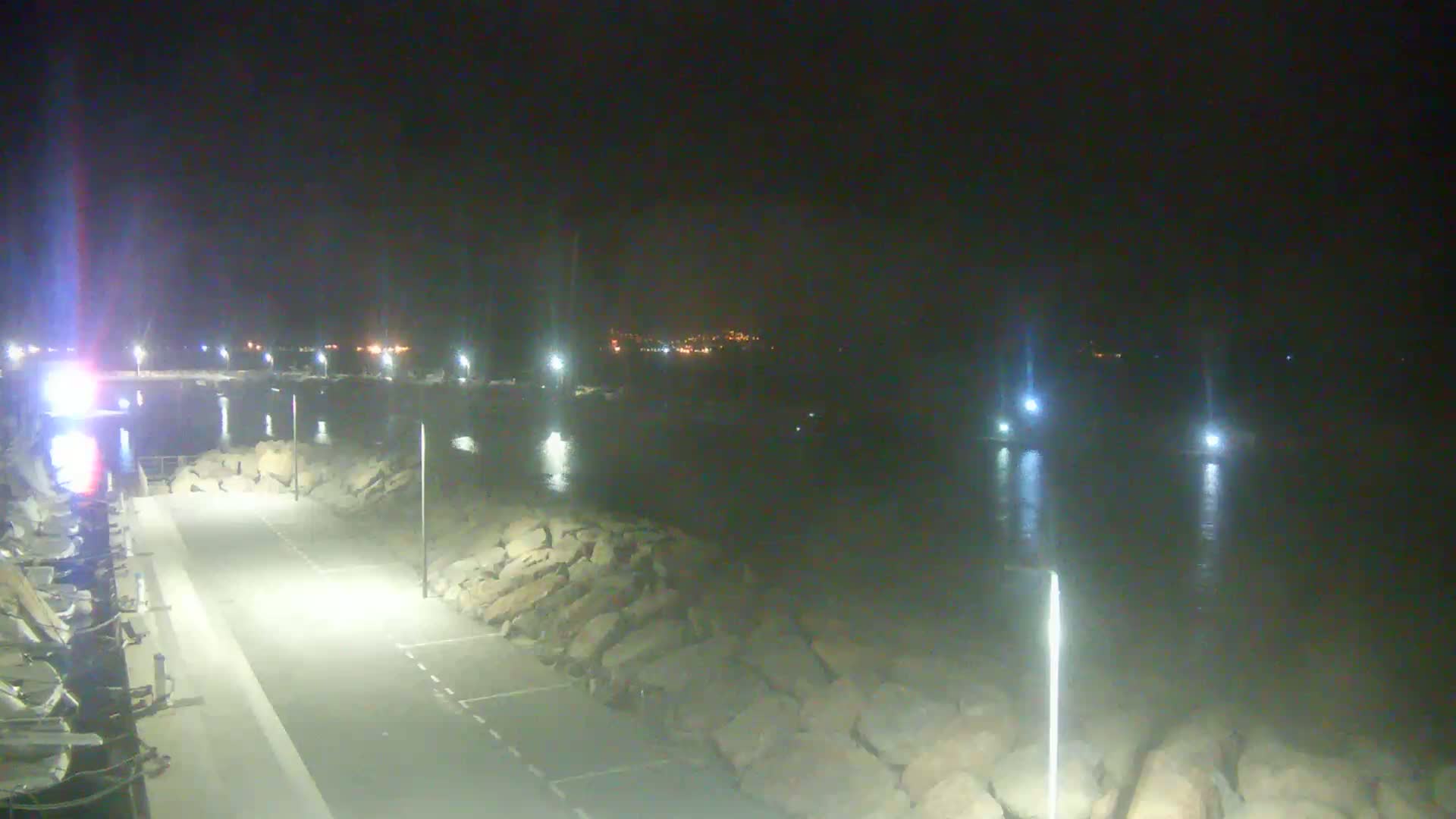 A nighttime view of a paved waterfront area with lights illuminating a rocky shoreline and calm water, with distant city lights visible across the water.