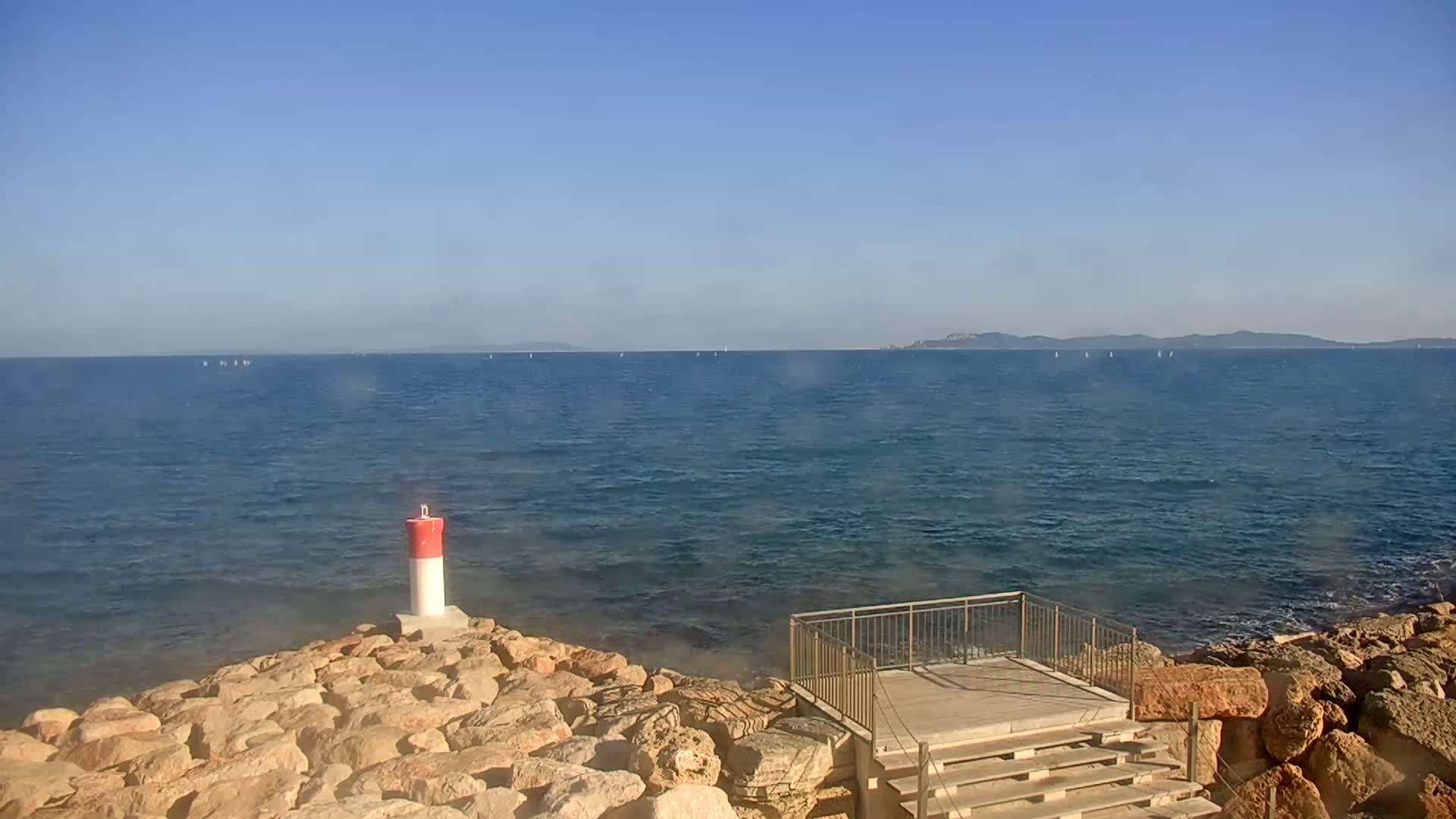 A rocky shoreline with a small wooden platform and steps overlooks a calm, blue sea with several sailboats visible in the distance under a clear, sunny sky.