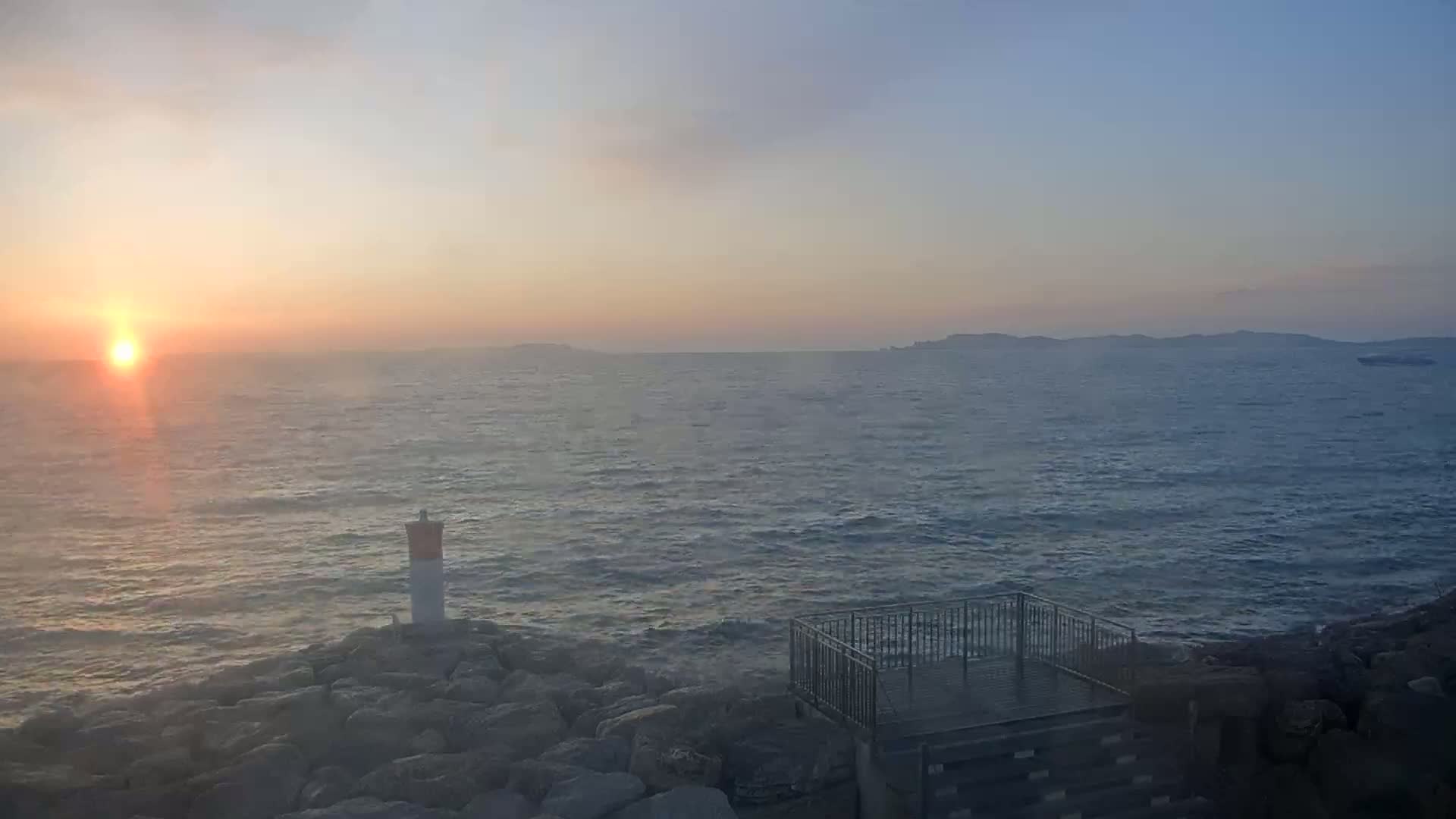 A rocky shoreline with a small viewing platform overlooks a calm sea at sunset, with distant islands visible on the horizon.