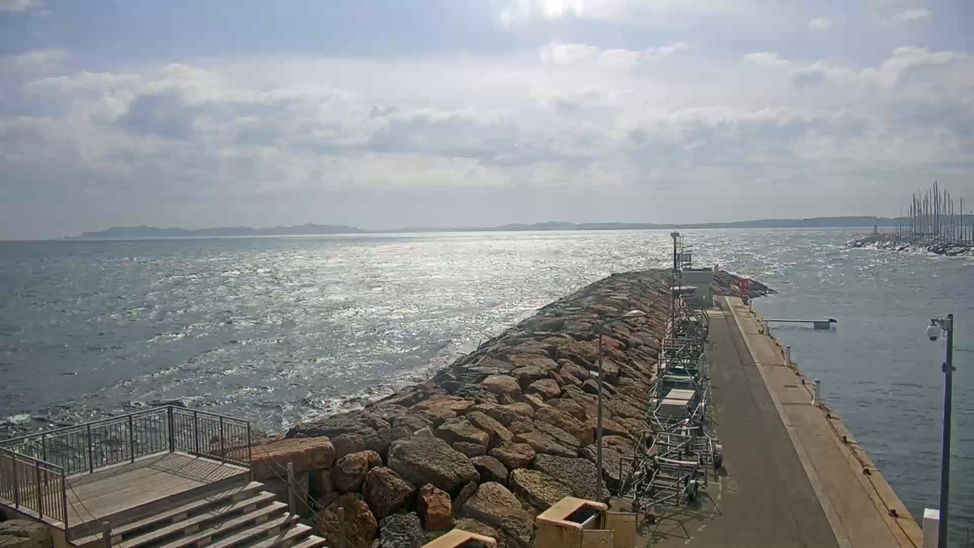 A rocky breakwater extends into a choppy sea under a partly cloudy sky, with a distant shoreline visible and a marina in the background.