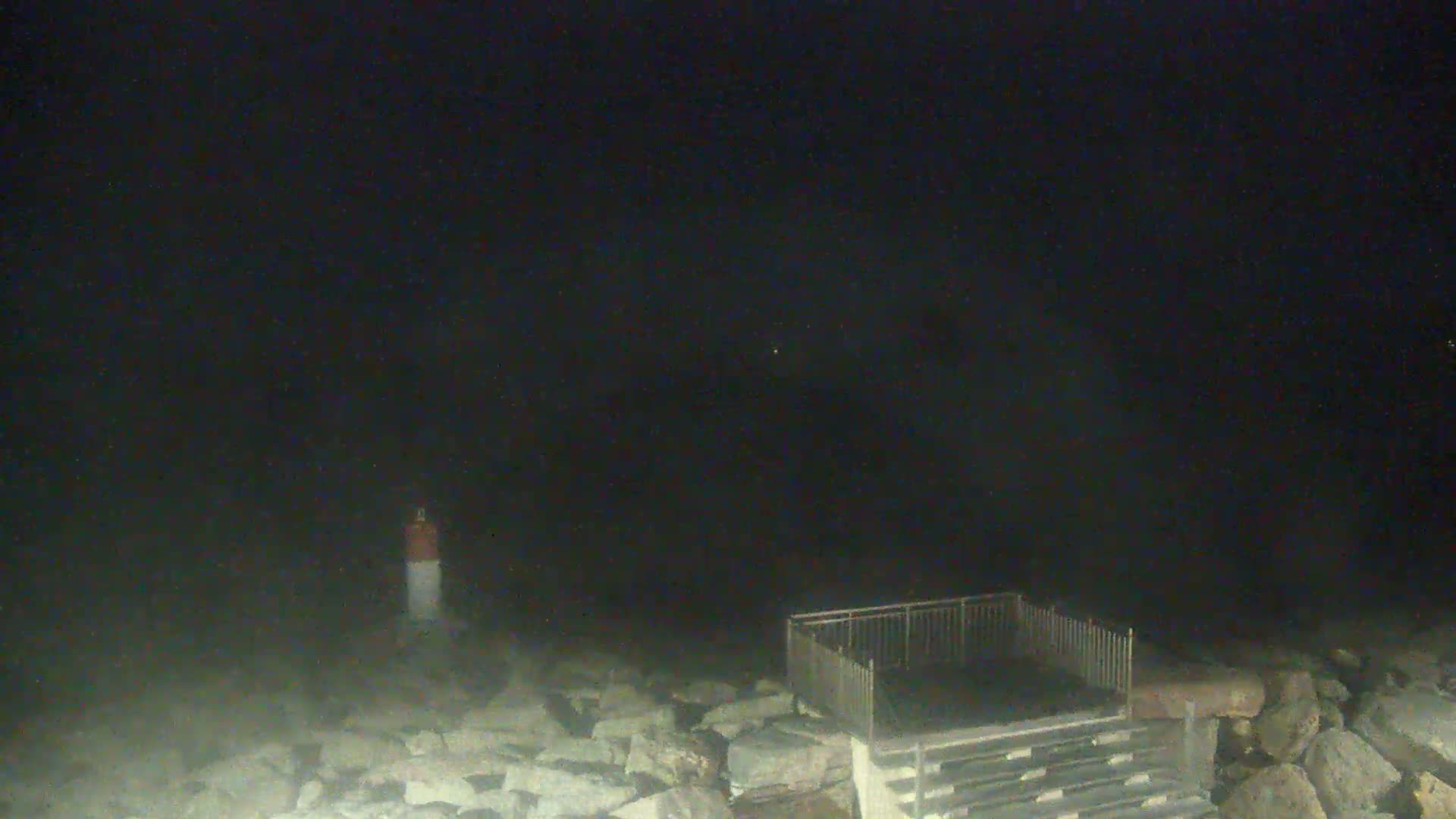 A nighttime scene shows a short, red and white post near a metal railing and stone steps leading down to a dark body of water under a dark sky.