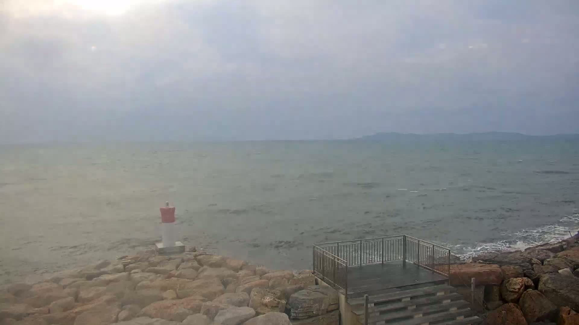 A hazy, overcast day shows a rocky shoreline with a small lighthouse and a viewing platform overlooking a choppy sea.