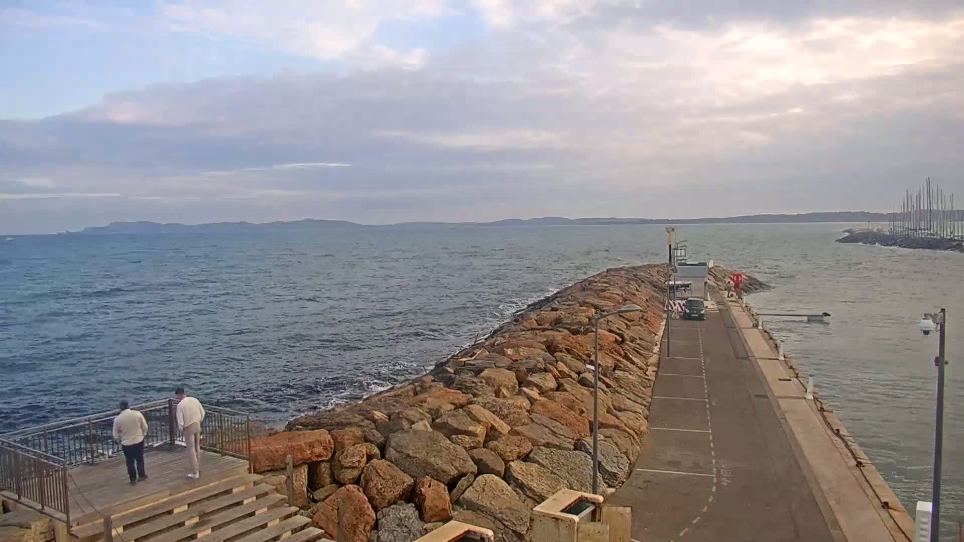 A rocky breakwater extends into a choppy sea under a cloudy sky, with two people standing on a viewing platform and a car parked on a nearby paved area.