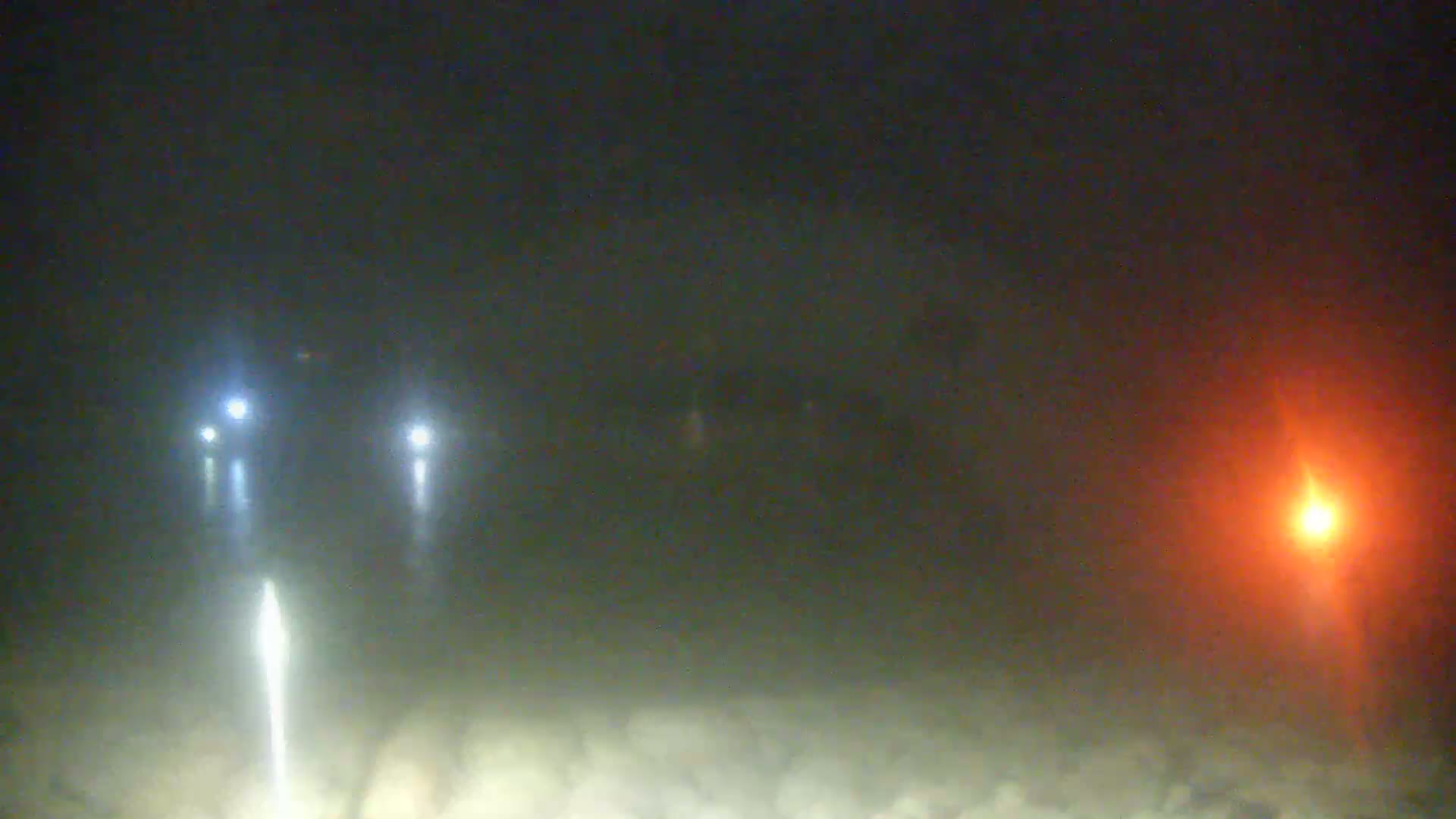 Two sets of vehicle headlights illuminate a foggy, nighttime scene, with a bright orange light visible in the distance to the right.