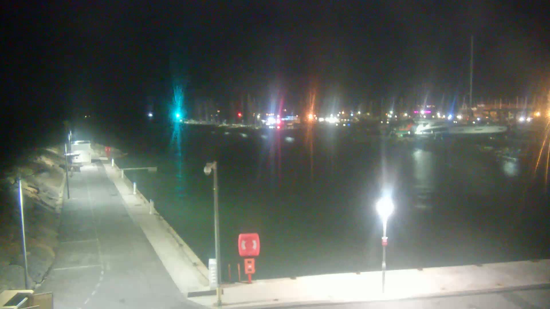A nighttime view of a harbor, showing a calm body of water with moored boats and a paved walkway with lights.