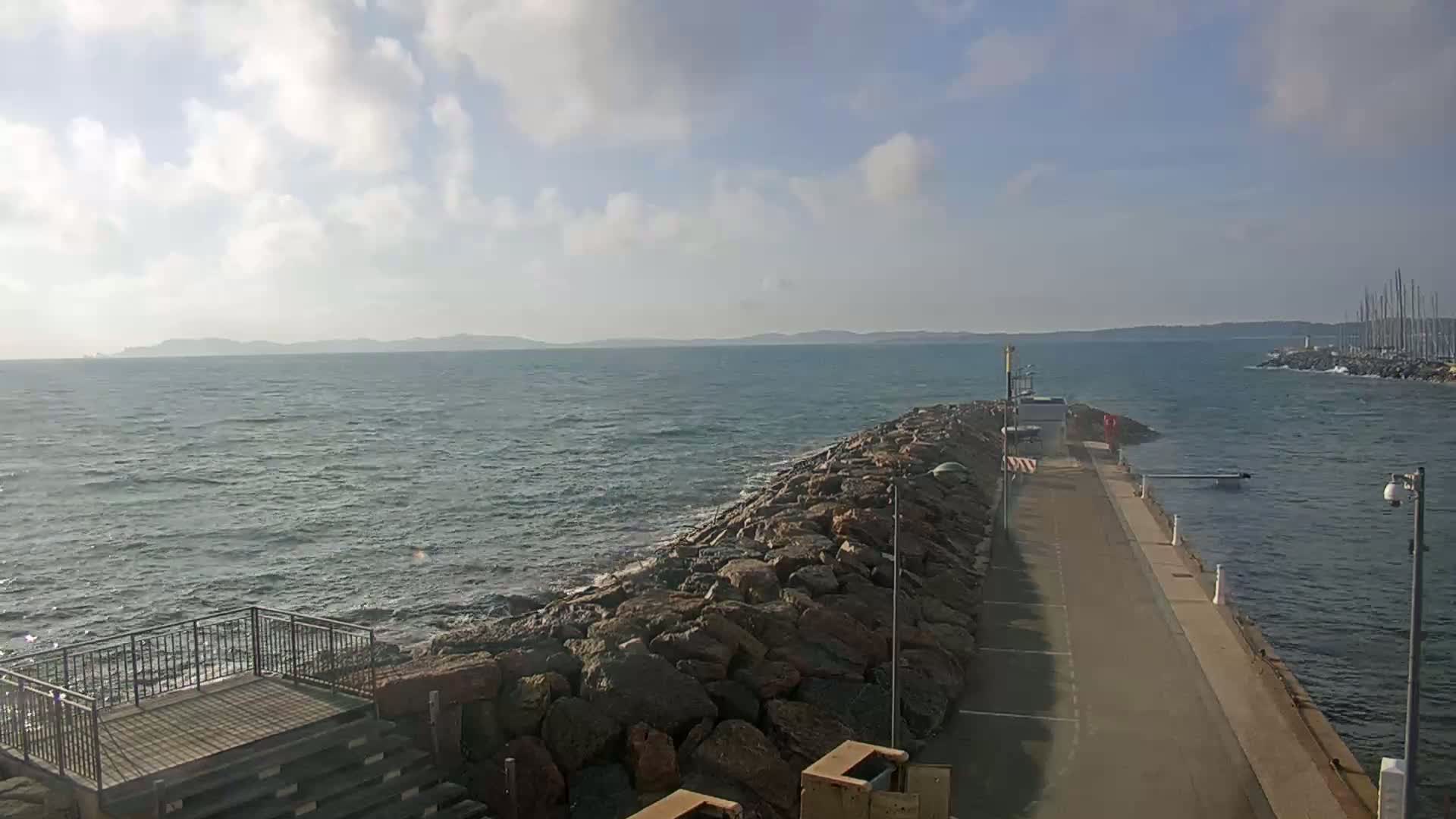 A rocky breakwater extends into a choppy sea under a partly cloudy sky, with land visible in the distance.