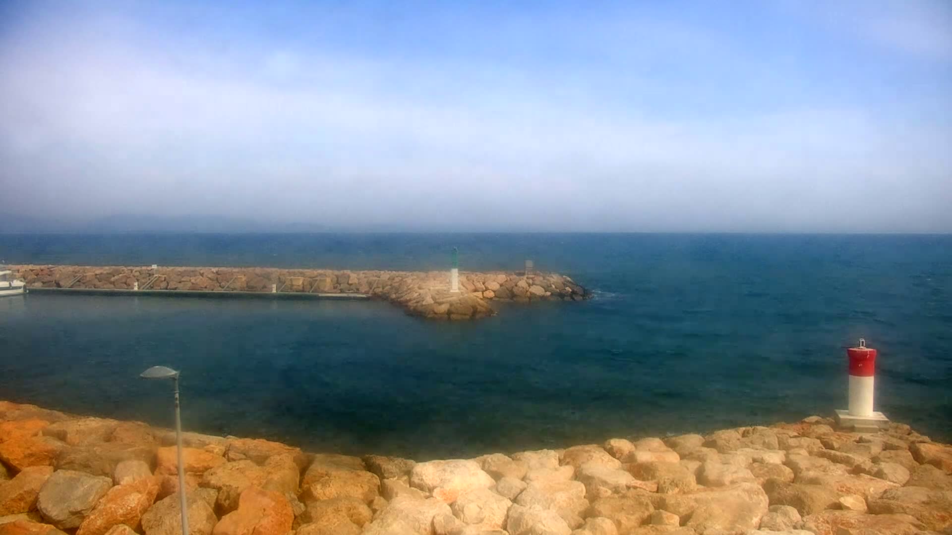 A calm, hazy day shows a rocky shoreline with a small harbor and a red and white navigational marker in the water.