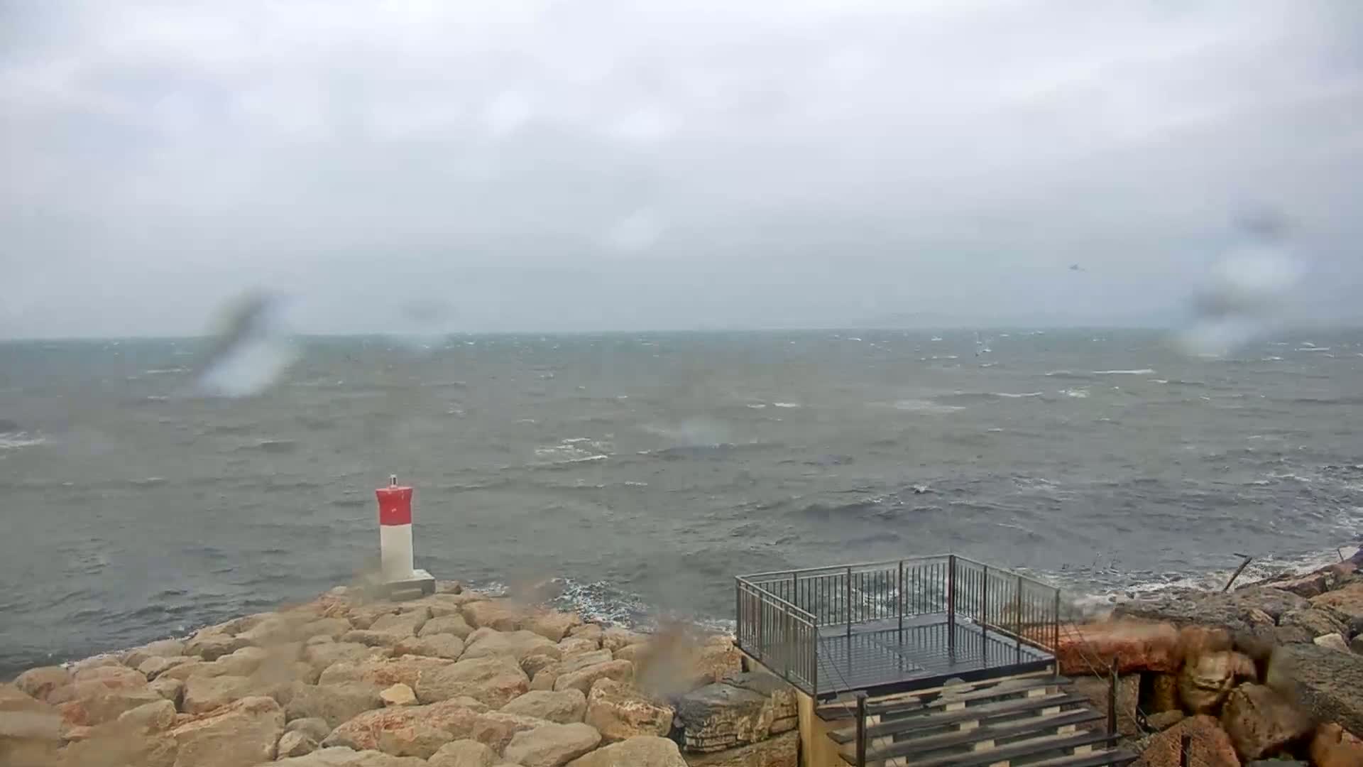A rocky shoreline with a small viewing platform and a navigational marker is shown during a windy, rainy day with choppy gray seas.