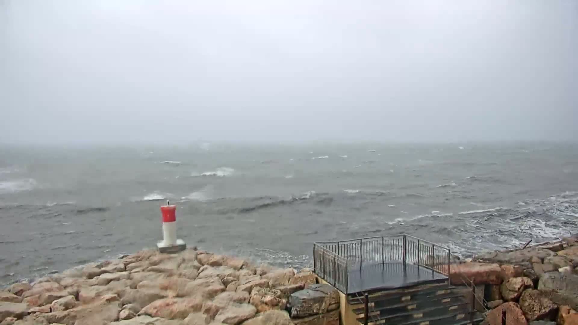 A rocky shoreline with a small red and white marker and a metal railing structure overlooks a choppy, gray sea under an overcast sky.