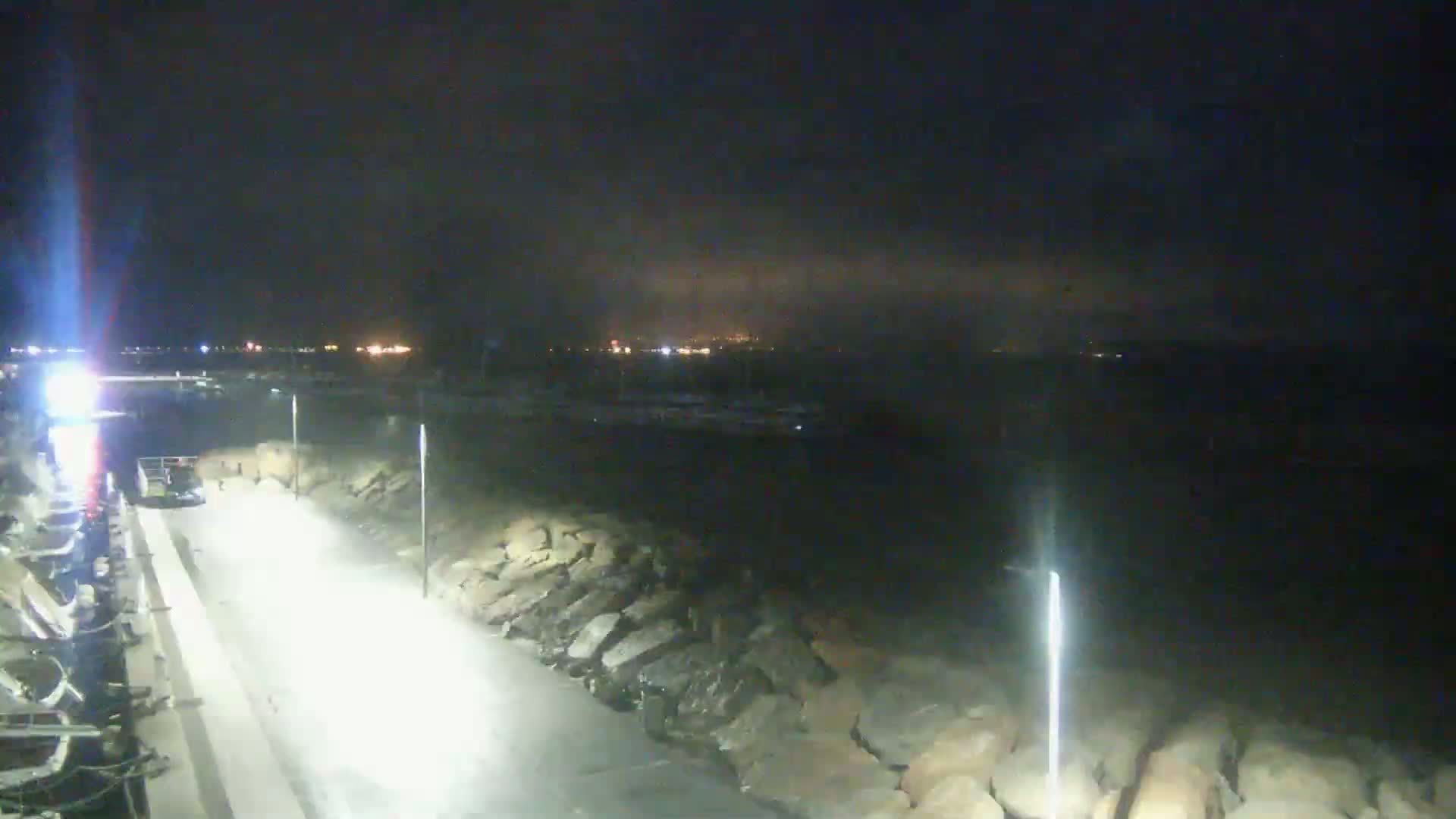 A nighttime view of a paved walkway beside a rocky shoreline, illuminated by bright lights, with a distant city skyline faintly visible under a dark, cloudy sky.