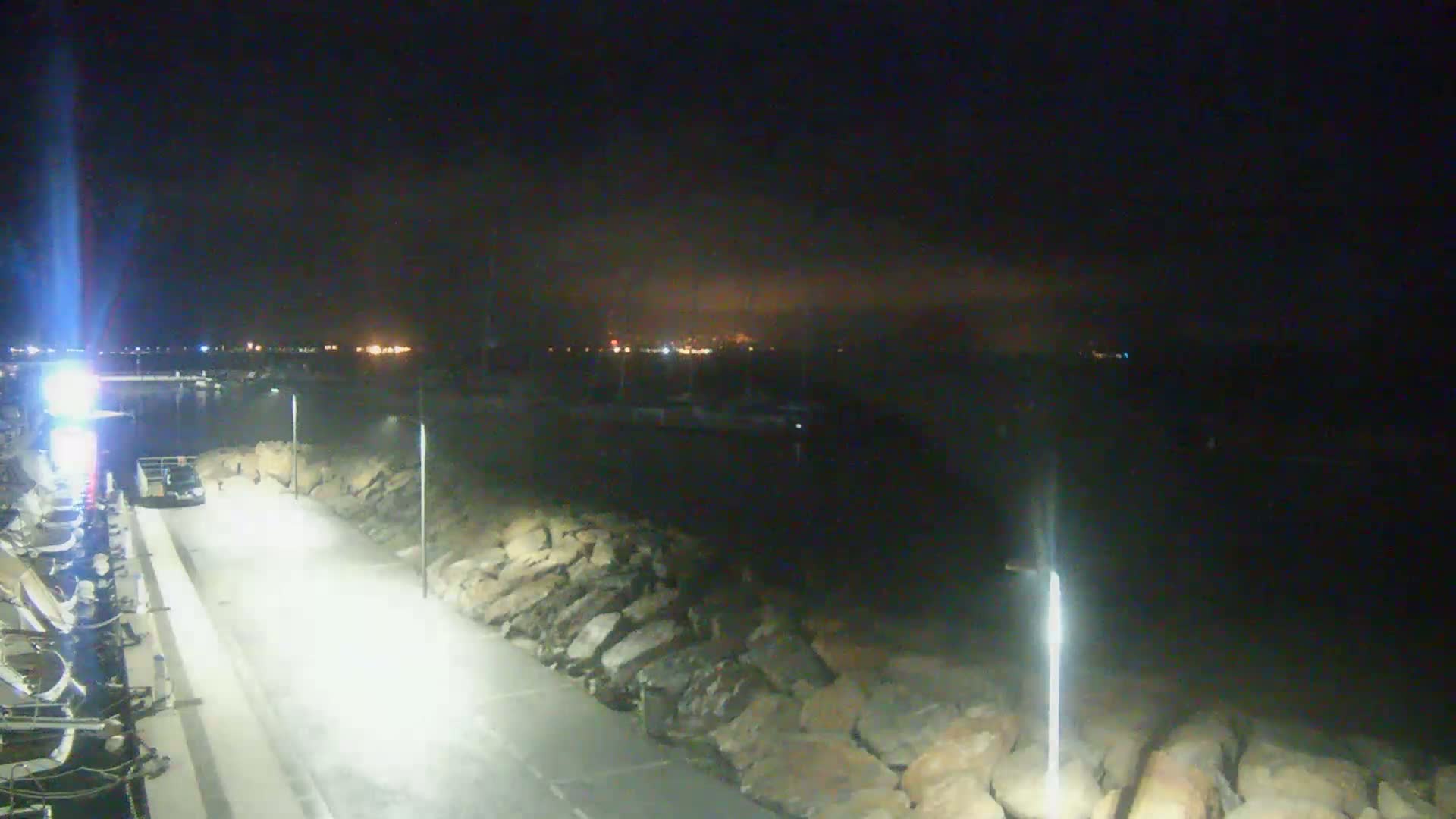 A nighttime view of a paved area next to a rocky shoreline, with a body of water and distant lights visible in the background under a dark, hazy sky.