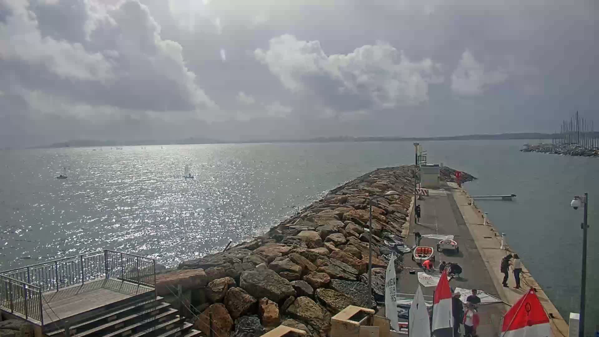 A rocky breakwater extends into a calm, sunlit body of water where several sailboats are visible under a partly cloudy sky.