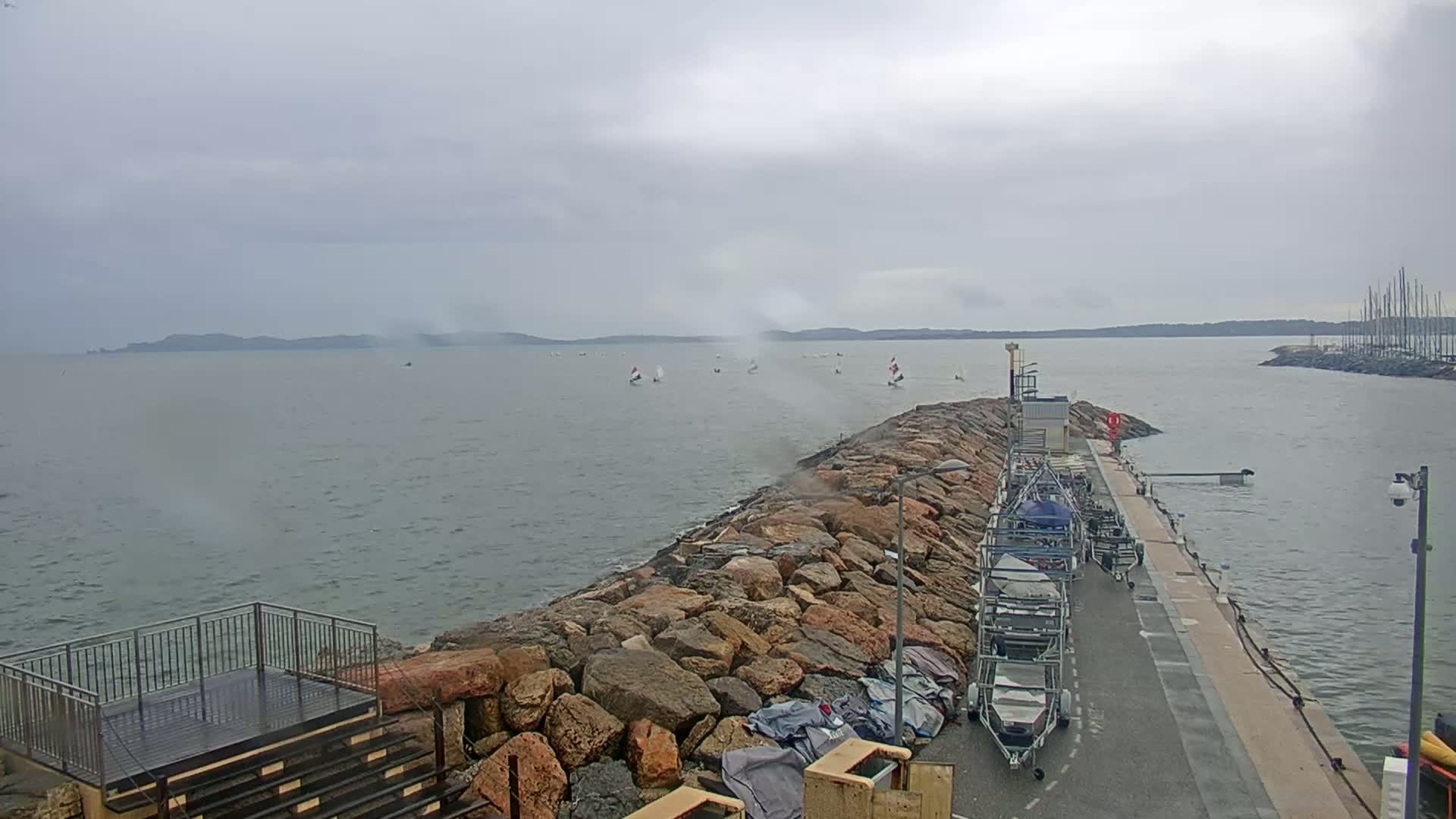 A rocky breakwater extends into a gray, choppy sea where several sailboats are visible under an overcast sky, with additional boats and boat storage visible along the shore.