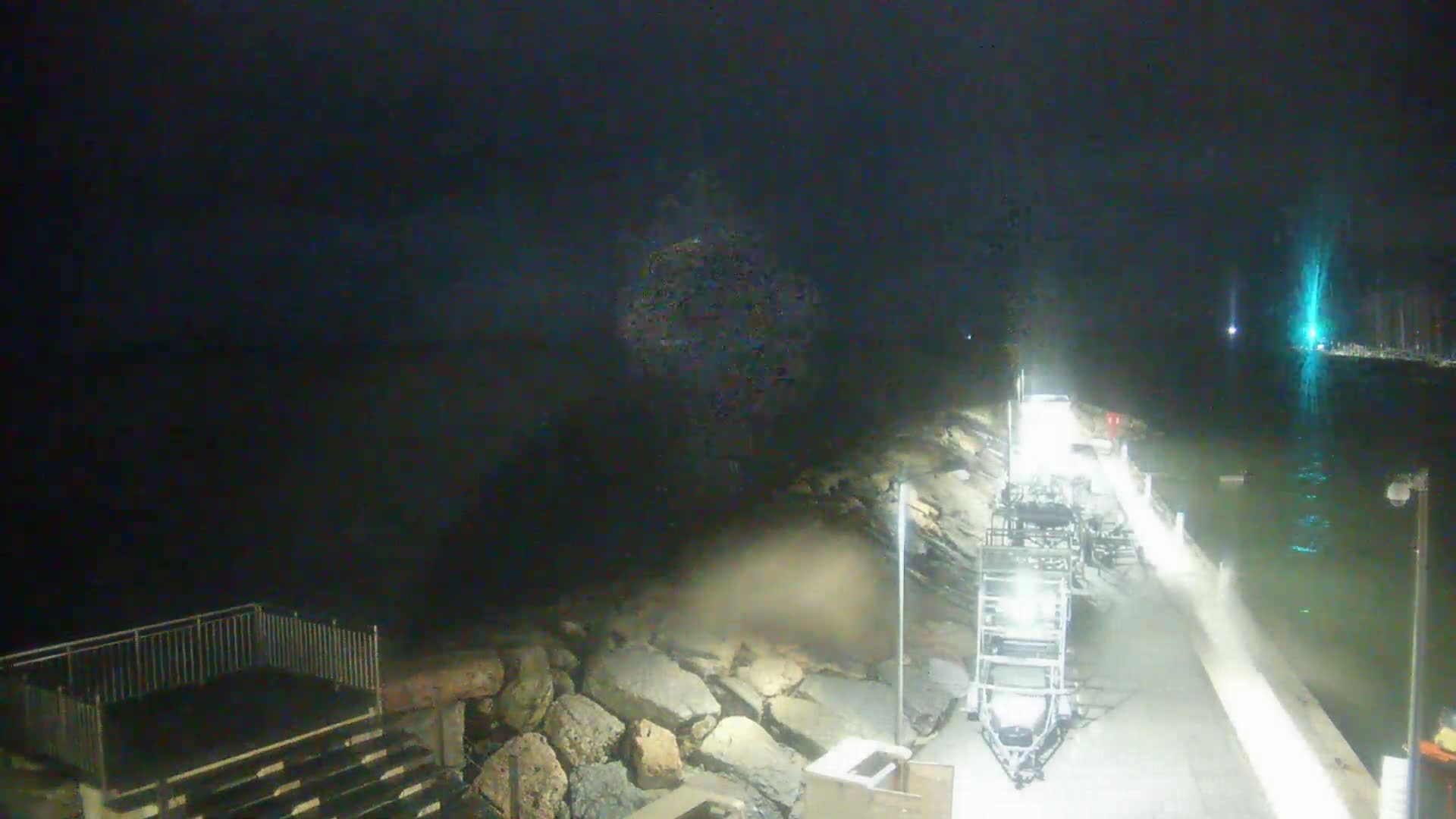 A nighttime view of a rocky waterfront, with a vehicle parked near a walkway, and dark, possibly stormy, water in the background.