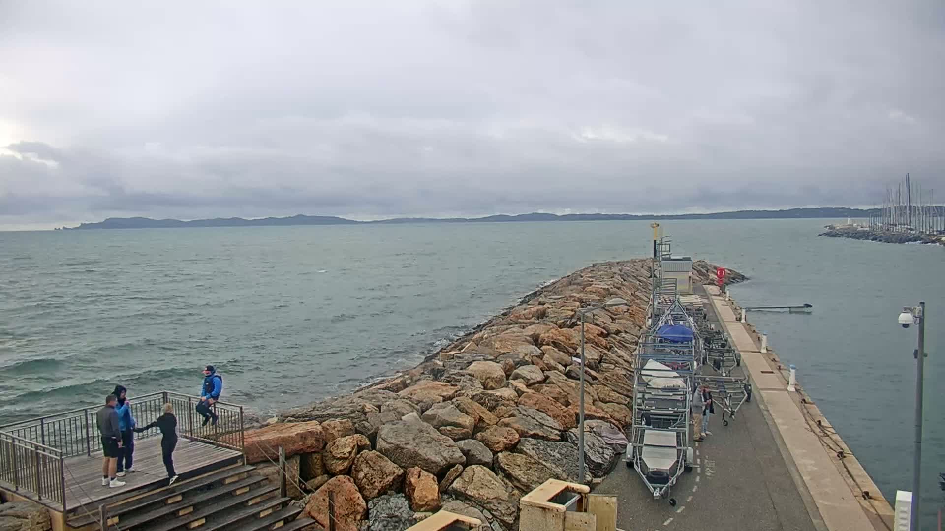 A rocky breakwater extends into a choppy sea under a cloudy sky, with several people standing on a wooden platform and several boats and boat trailers parked nearby.