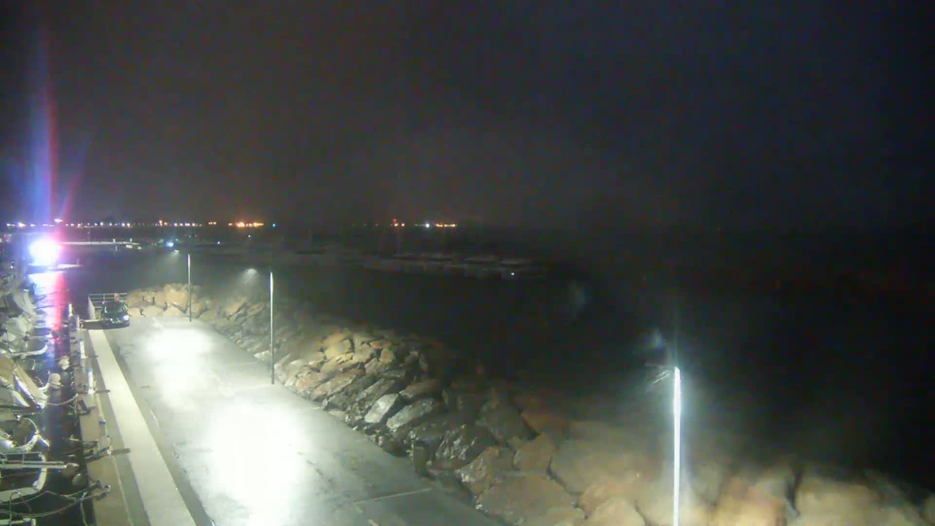 A nighttime view shows a dark, possibly stormy, ocean next to a paved area with lights, lined with rocks and parked boats.