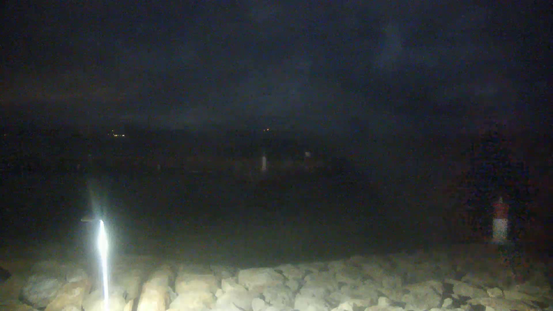 A nighttime view of a rocky shoreline with a bright light in the foreground, under a dark, cloudy sky.