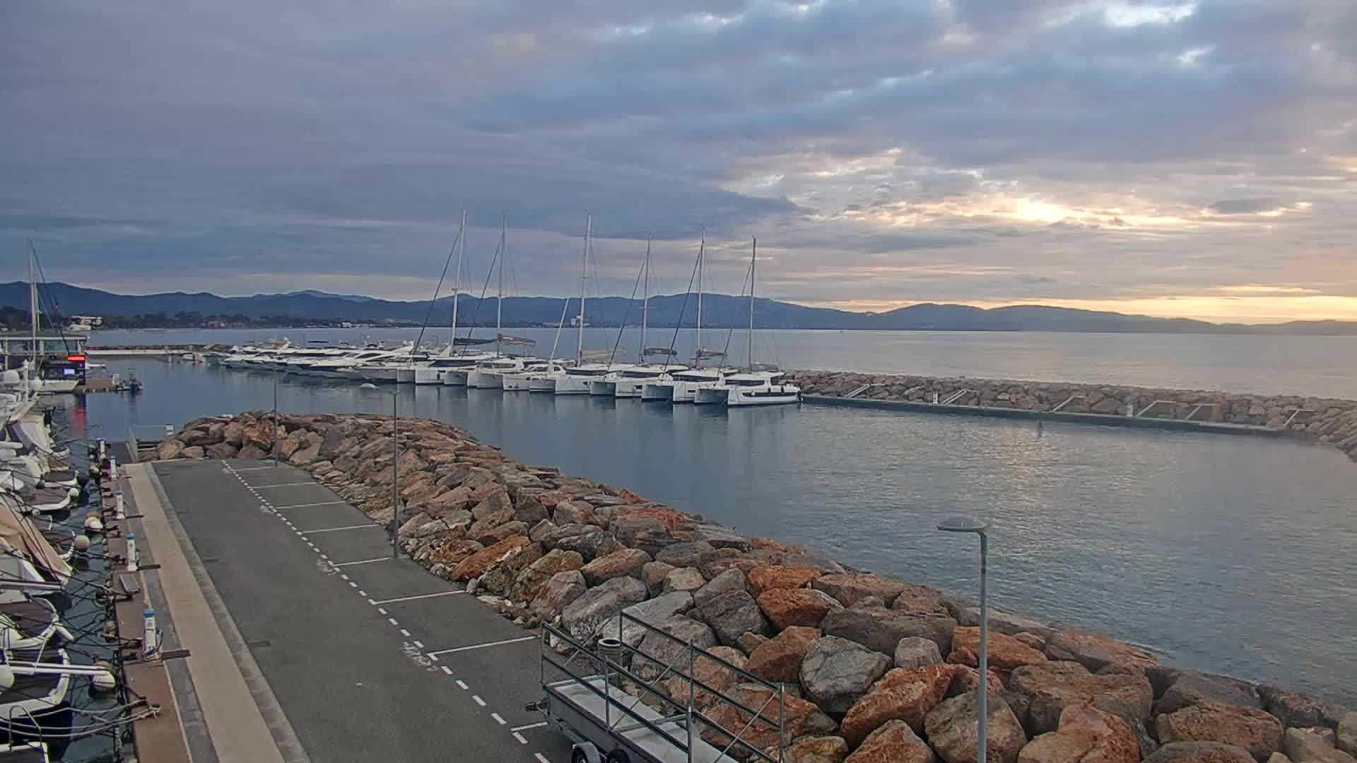 A calm harbor at sunrise or sunset, filled with several sailboats and yachts moored alongside a rocky breakwater.