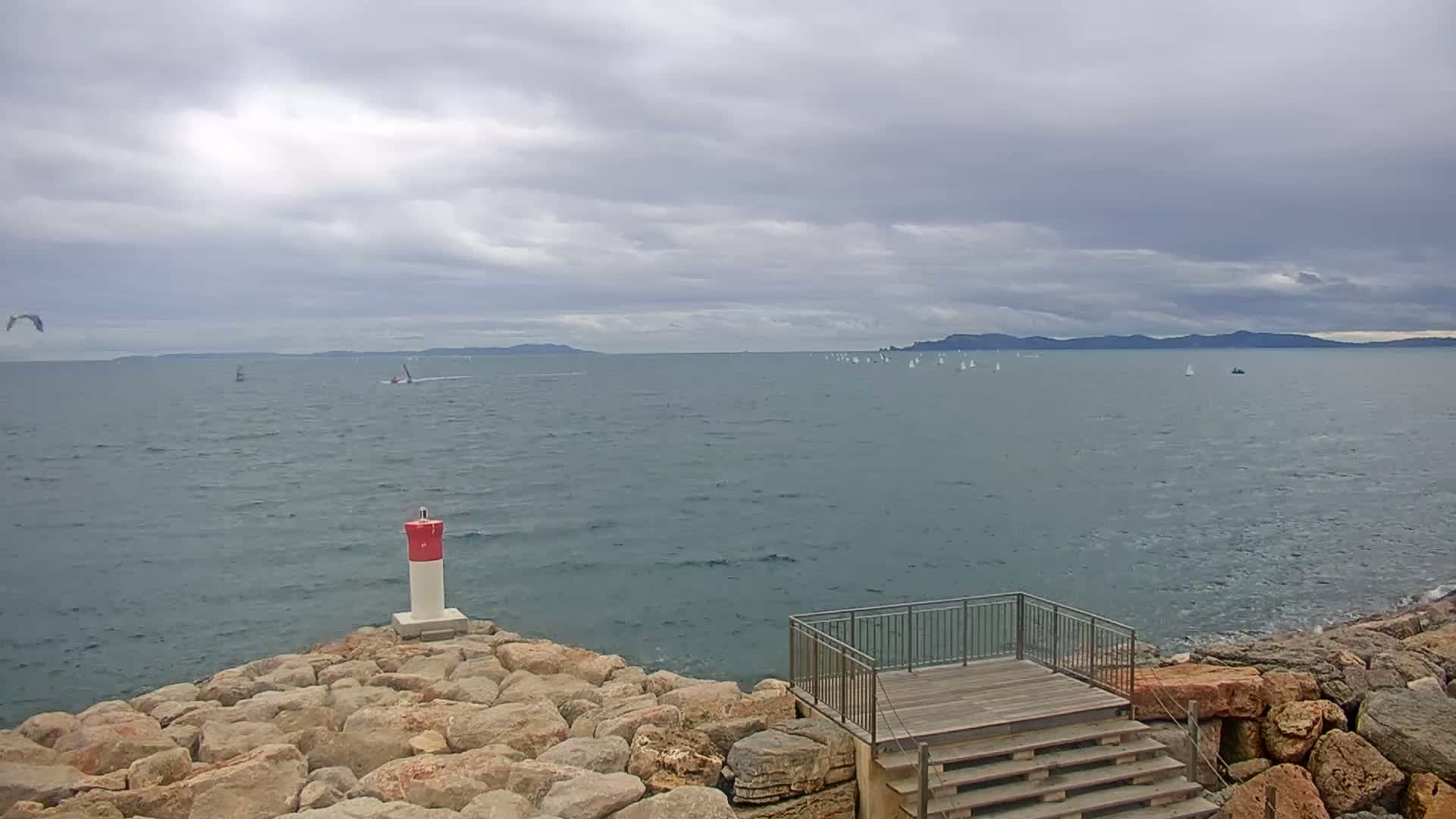 A rocky shoreline with a small lighthouse and wooden steps overlooks a calm sea with numerous sailboats and a distant coastline under a cloudy sky.