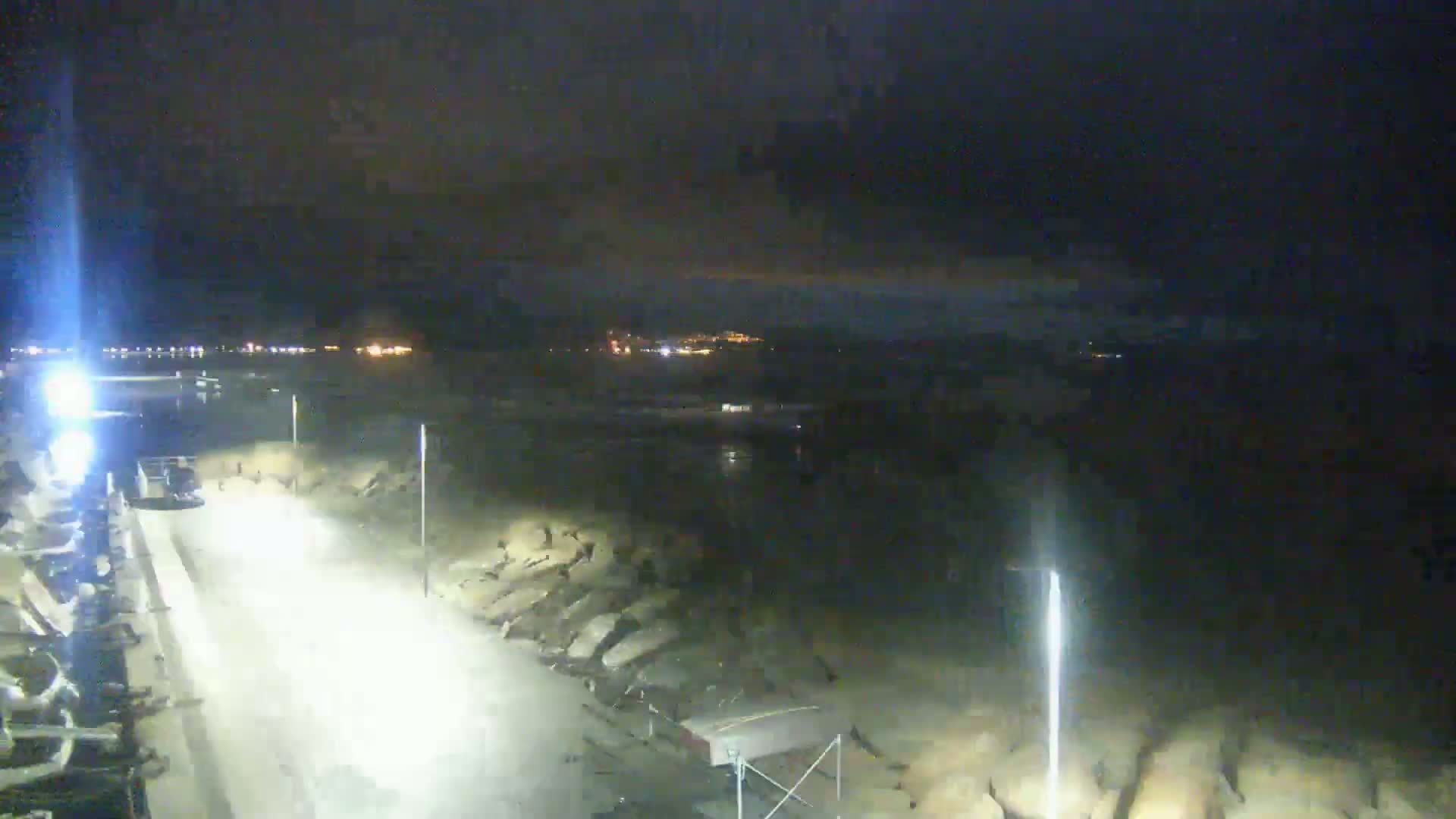 A nighttime view of a dark, possibly overcast, shoreline with a brightly lit roadway and distant lights across the water.