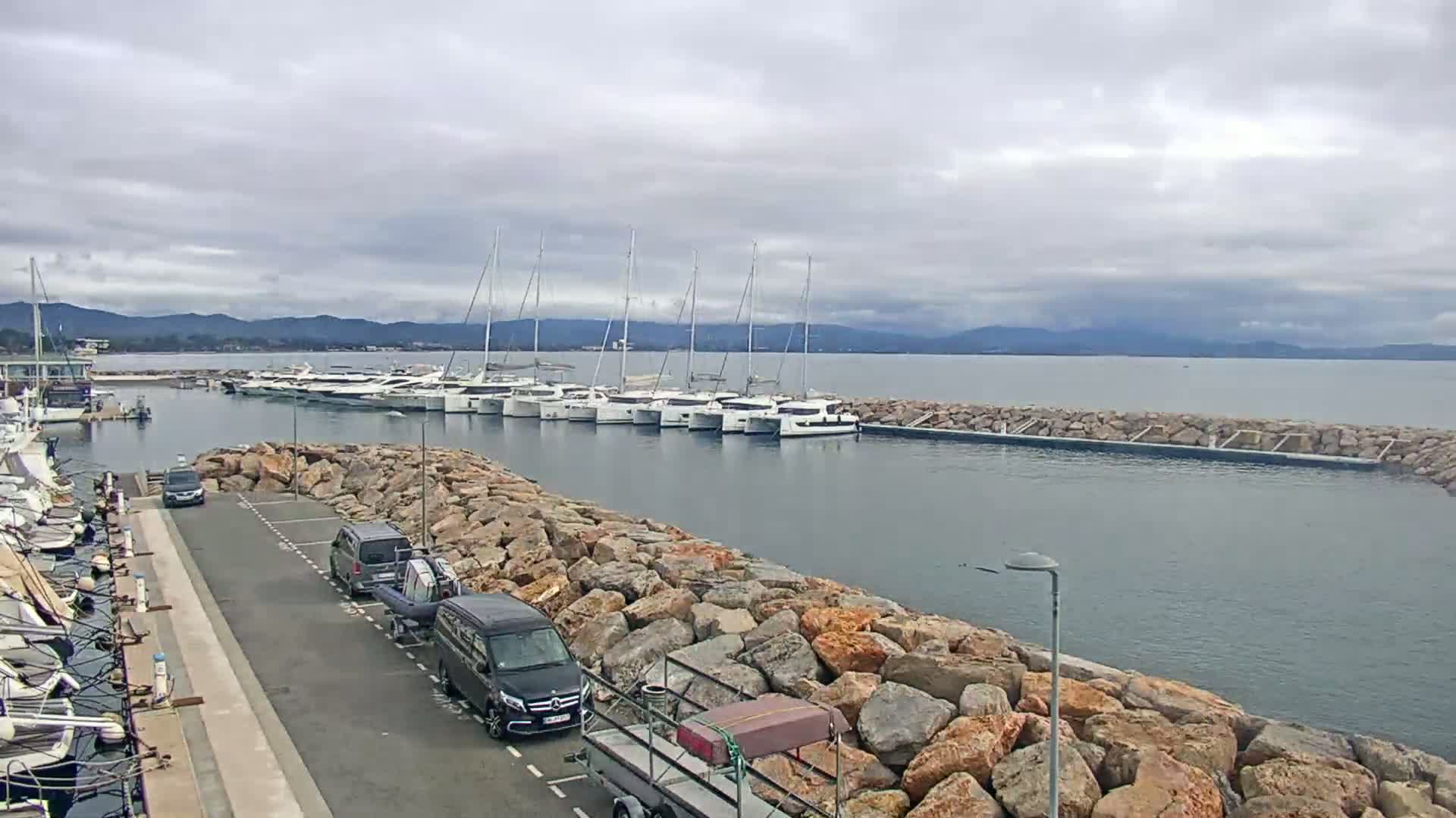 A marina with numerous sailboats and yachts is visible under an overcast sky, bordered by a rocky breakwater with several cars parked nearby.