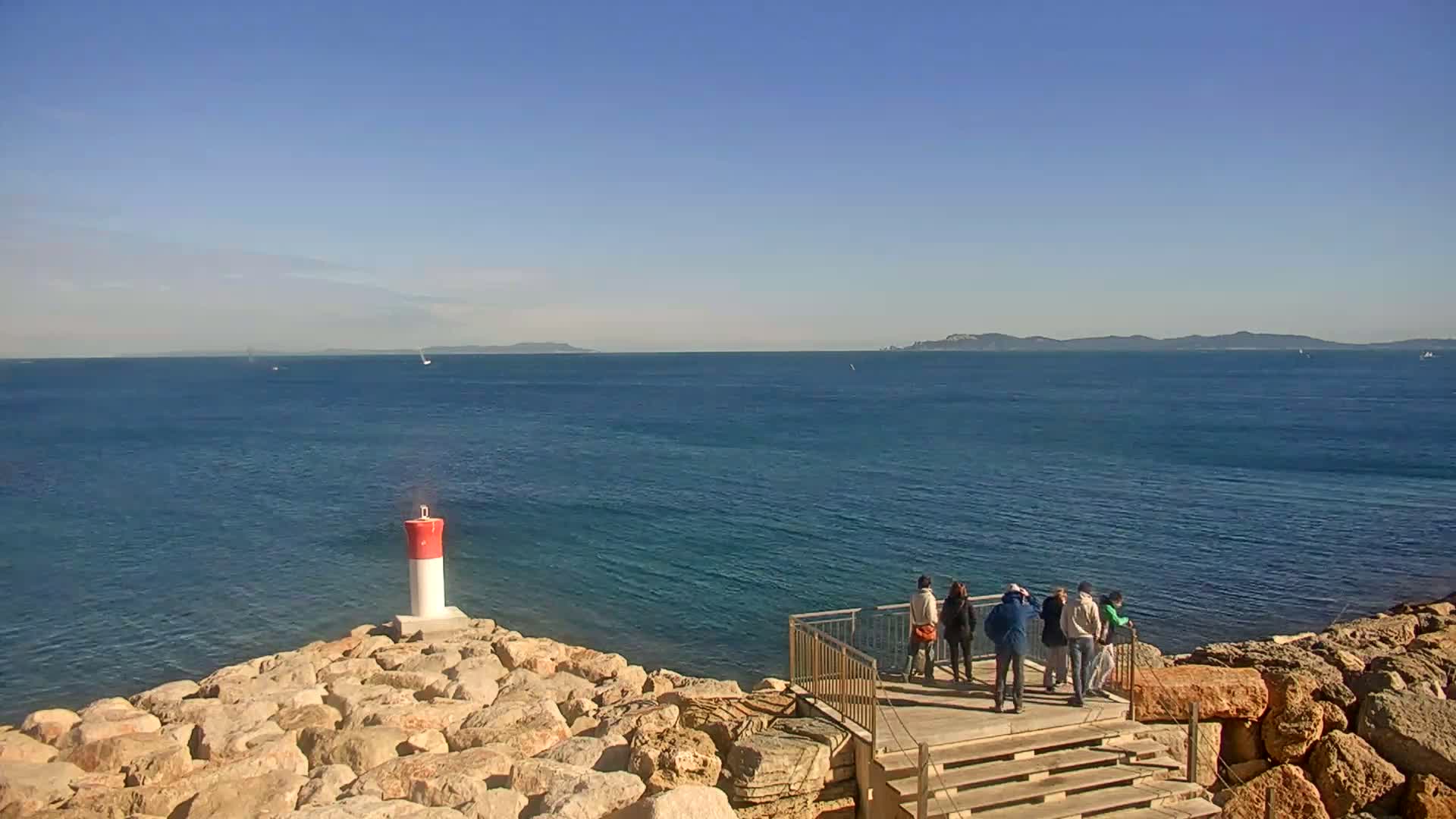 A group of people stand on a wooden platform overlooking a calm blue sea, on a sunny day, with a small red and white lighthouse on a rocky outcropping nearby.