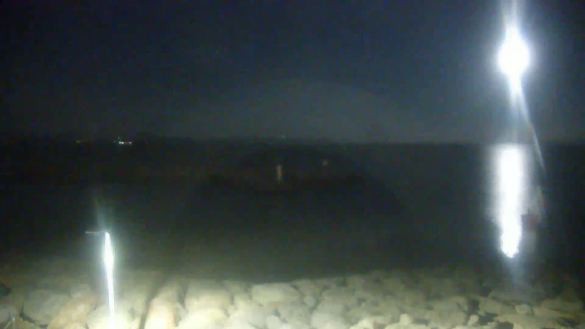 A nighttime, low-light view of a rocky shoreline with a body of dark water extending to a distant horizon under a dark sky, illuminated by bright light sources in the foreground and distance.