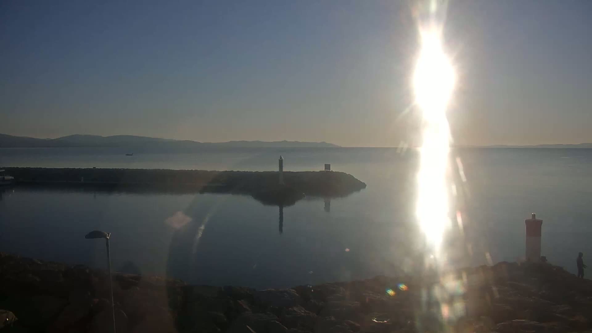 A bright sun shines over a calm body of water, with a small island featuring a lighthouse visible in the distance and a rocky shoreline in the foreground.