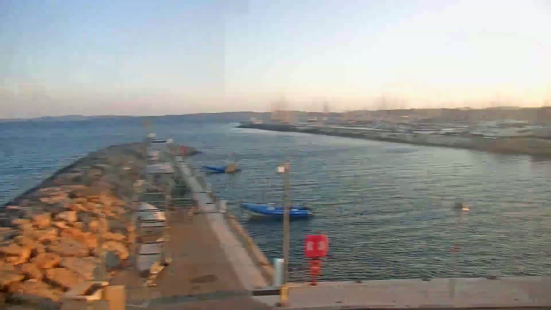 A calm, clear day reveals a harbor with a few small boats docked near a rocky breakwater, leading to a larger body of water and a distant town.